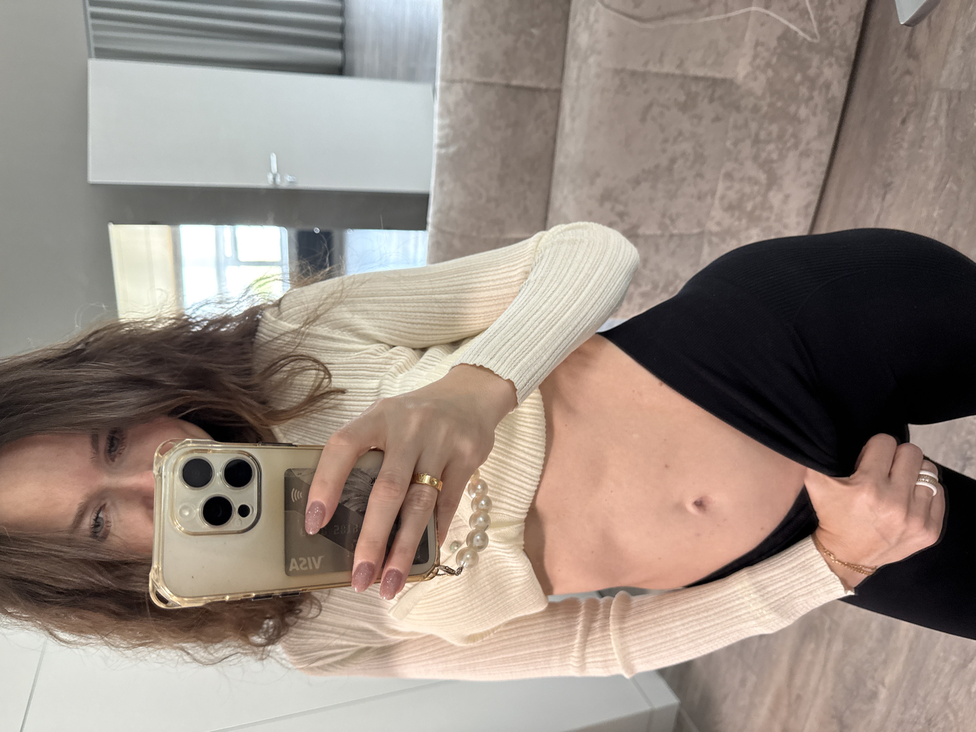 A woman is taking a selfie in a room, likely in an apartment. She is holding an iPhone with a case that appears to have a credit card inserted in it. She is wearing a cream-colored, long-sleeved ribbed sweater and black leggings, which she is pulling up slightly to reveal her stomach. There is a sofa visible in the background. The location in the city of Khimki, Russia, is not discernible from the image.