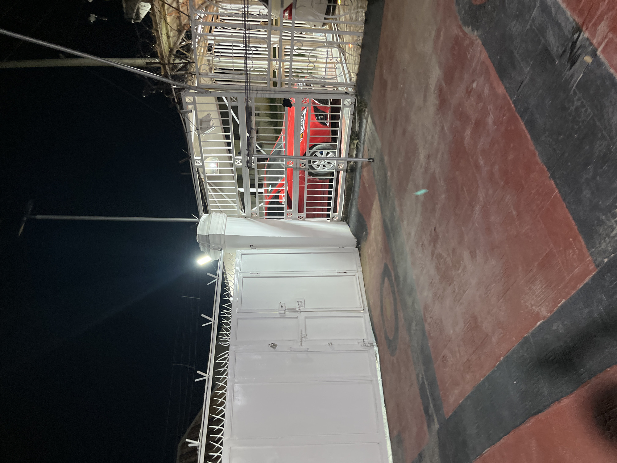 The image depicts a night scene at a gate in Abuja, Nigeria. The gate is white and made of metal, with a locked padlock visible. Beyond the gate, a red car is parked, partially obscured by the fencing. The ground in front of the gate is paved with red and black sections, possibly a driveway or courtyard. The lighting suggests artificial illumination from streetlights or security lights. The overall impression is of a secure residential or commercial property entrance at night.