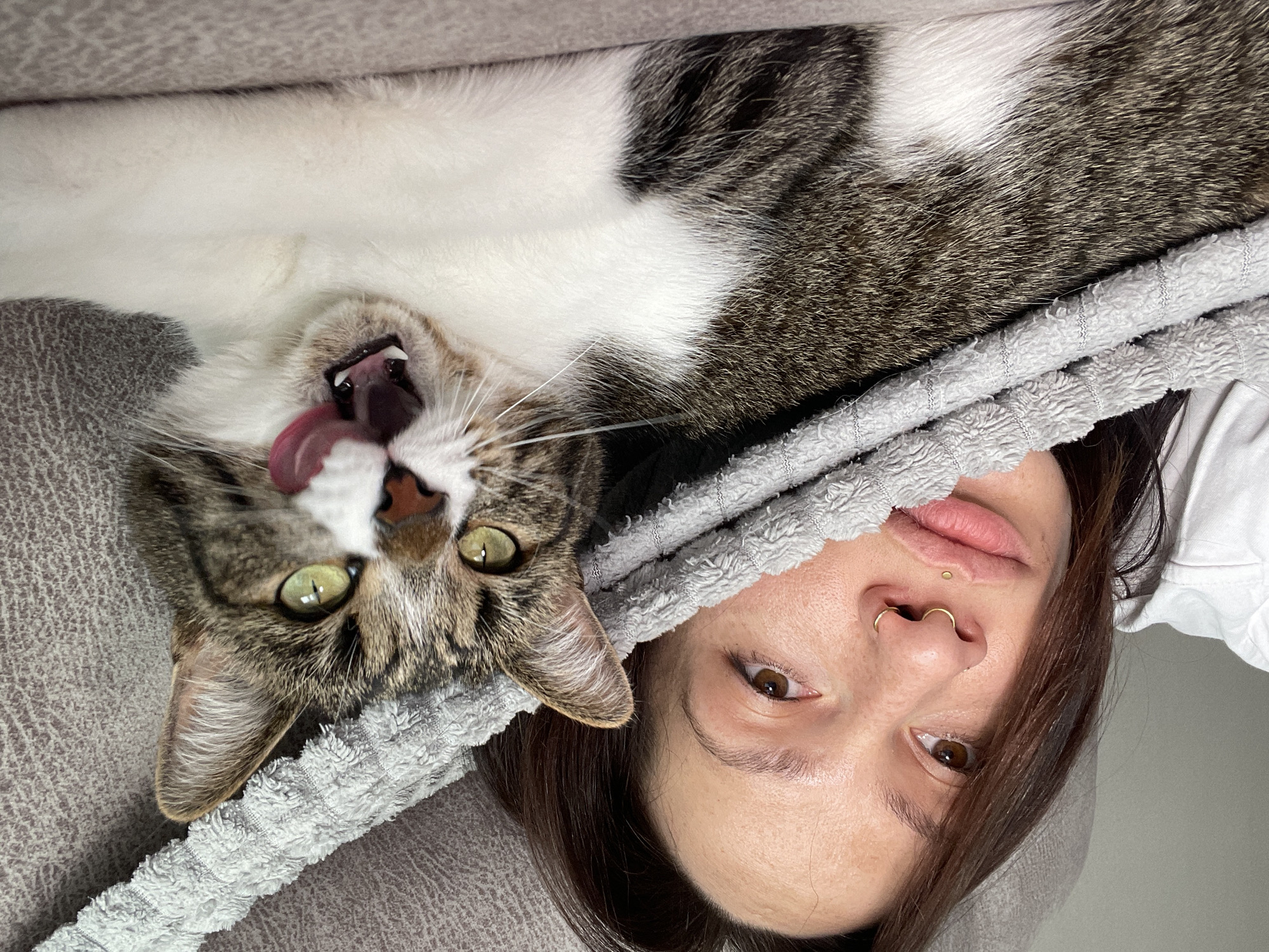 A woman and a cat are lying down next to each other. The cat is on the left, and the woman is on the right. They are both facing the camera. The cat is lying on its back, and its tongue is sticking out. The woman is looking at the camera. A gray towel is covering part of the woman's face. The location is not identifiable as Pervomaysky, Russia from the image.