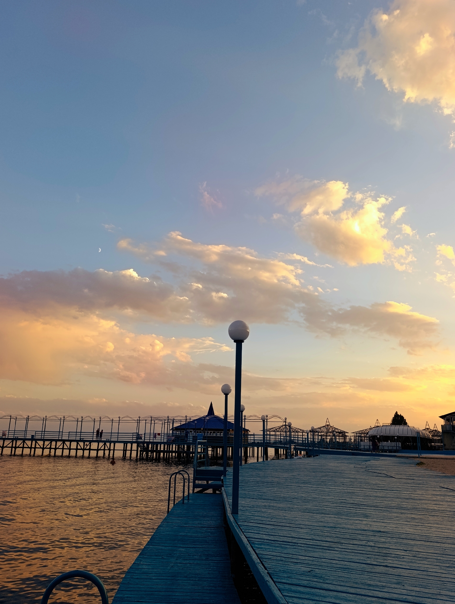 The image depicts a wooden pier extending into a body of water under a twilight sky. The sky is filled with wispy clouds, some tinged with orange and yellow from the setting sun. A crescent moon is visible in the upper left quadrant of the sky. The pier is made of weathered blue-gray wooden planks and is lined with lampposts topped with spherical lights. In the distance, a larger structure with a blue roof, possibly a pavilion or restaurant, is situated on the pier. Fencing runs along the sides of the pier, with figures of two people visible walking on it in the mid-ground. The water is calm, reflecting the colors of the sky. To the right of the pier, a sandy area with some covered structures can be seen. The scene is set in Bayet, Kyrgyzstan.