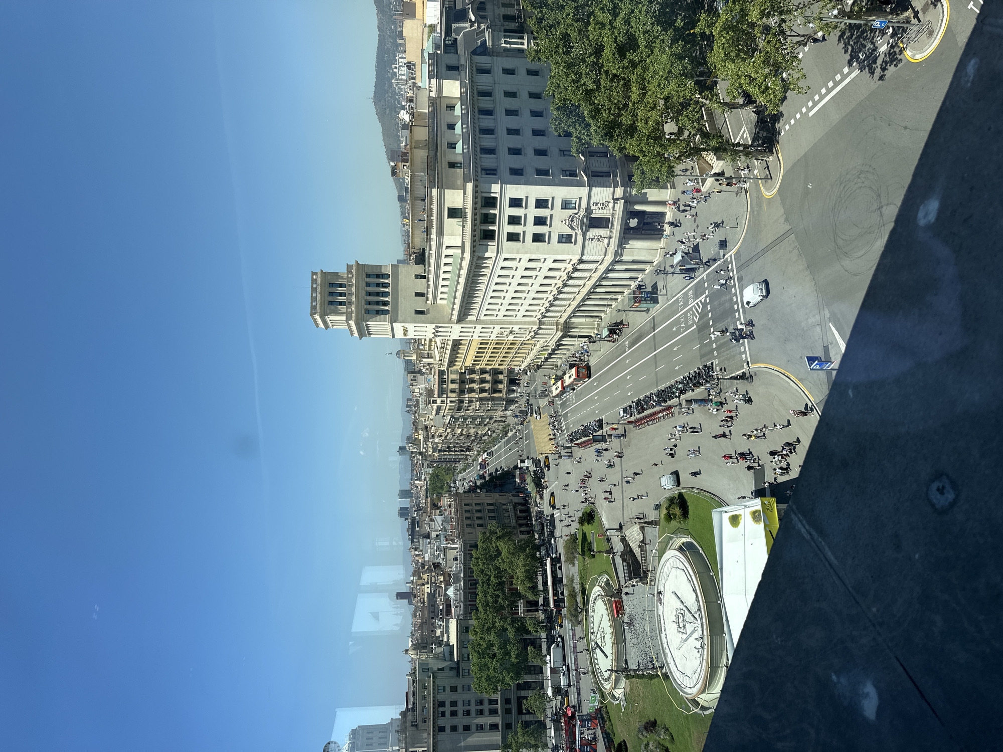 This image captures a wide-angle, elevated view of Plaça de Catalunya in Barcelona, Spain, on a clear, sunny day. The perspective is from a window, indicated by reflections on the glass.
The foreground shows a dark, textured surface, likely a window sill or balcony. Dominating the mid-ground is Plaça de Catalunya, a large public square with several circular fountains, one of which has a central sculptural element. Lush green grass surrounds the fountains, and numerous pigeons are visible in and around them.
The square is bustling with activity. Pedestrians are walking in various directions, some congregating near the fountains, others crossing the streets. A significant amount of traffic is present on the wide avenues radiating from the square. Numerous cars, buses (including red double-decker tourist buses), taxis, and motorcycles are on the road. A white car is seen driving through an intersection, and a line of parked motorcycles is visible along one side of the street.
Several prominent buildings surround the square. To the right, a large, light-colored building with a distinctive tower stands prominently. An Apple Store logo is visible on the facade of a building on the corner. To the left, other multi-story buildings, characteristic of European city architecture, line the streets. In the distance, the cityscape of Barcelona stretches out, with more buildings and a hazy view of hills or mountains. The sky is a clear, bright blue, with a few faint streaks that could be contrails or atmospheric phenomena.