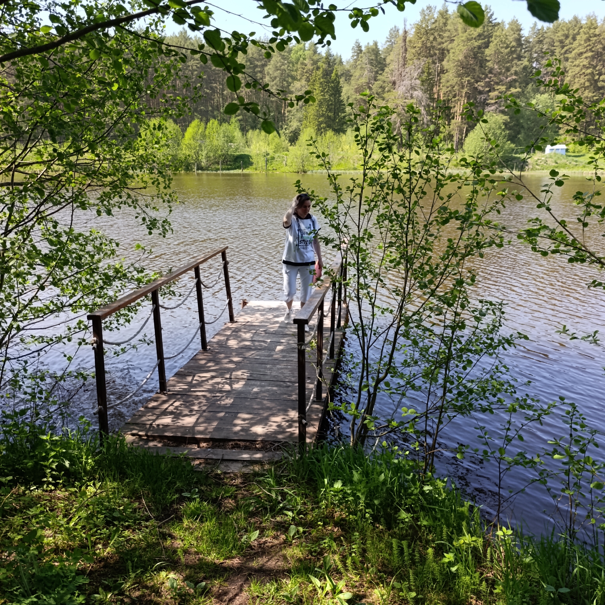 The image shows a woman standing on a wooden pier extending into a body of water. The pier has metal railings with rope accents. The water appears calm with slight ripples. The pier is bordered by green vegetation, including grass and leafy bushes. In the background, a forest of green trees is visible. The image was taken in the city of Prikamskii, Russia.