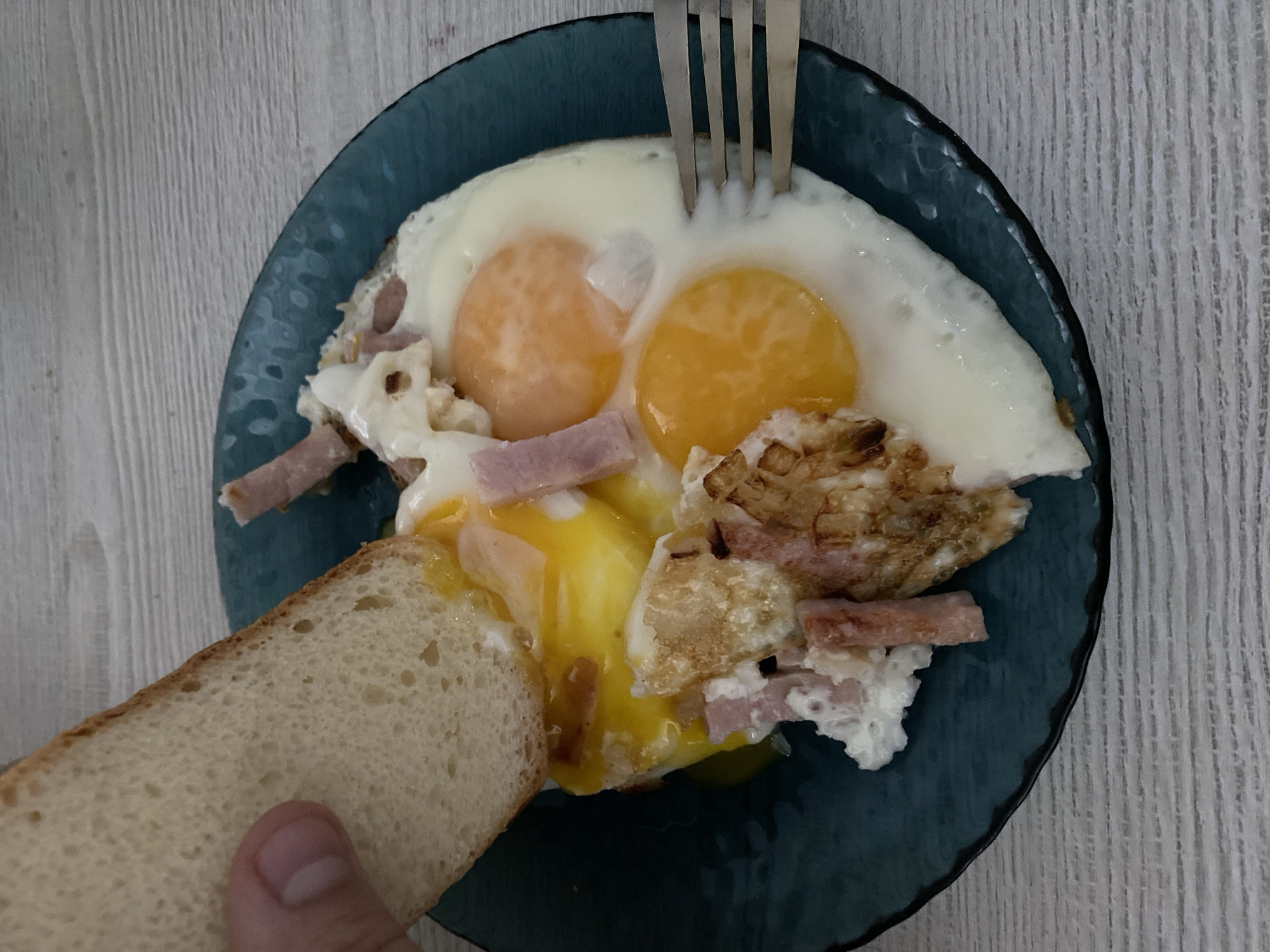 A plate of eggs, ham and bread is viewed from directly above. The dish is a dark blue textured ceramic plate. Two sunny-side up eggs occupy the upper half of the plate. A silver metal fork is sticking into the eggs near the top center of the plate. Bits of ham and cooked egg whites surround the two eggs. A slice of bread is being held to soak up the runny yolk of the egg on the lower left side of the plate. The background is a light gray wood-like surface.
There is no information available to identify the location as Chaikovsky, Russia.
