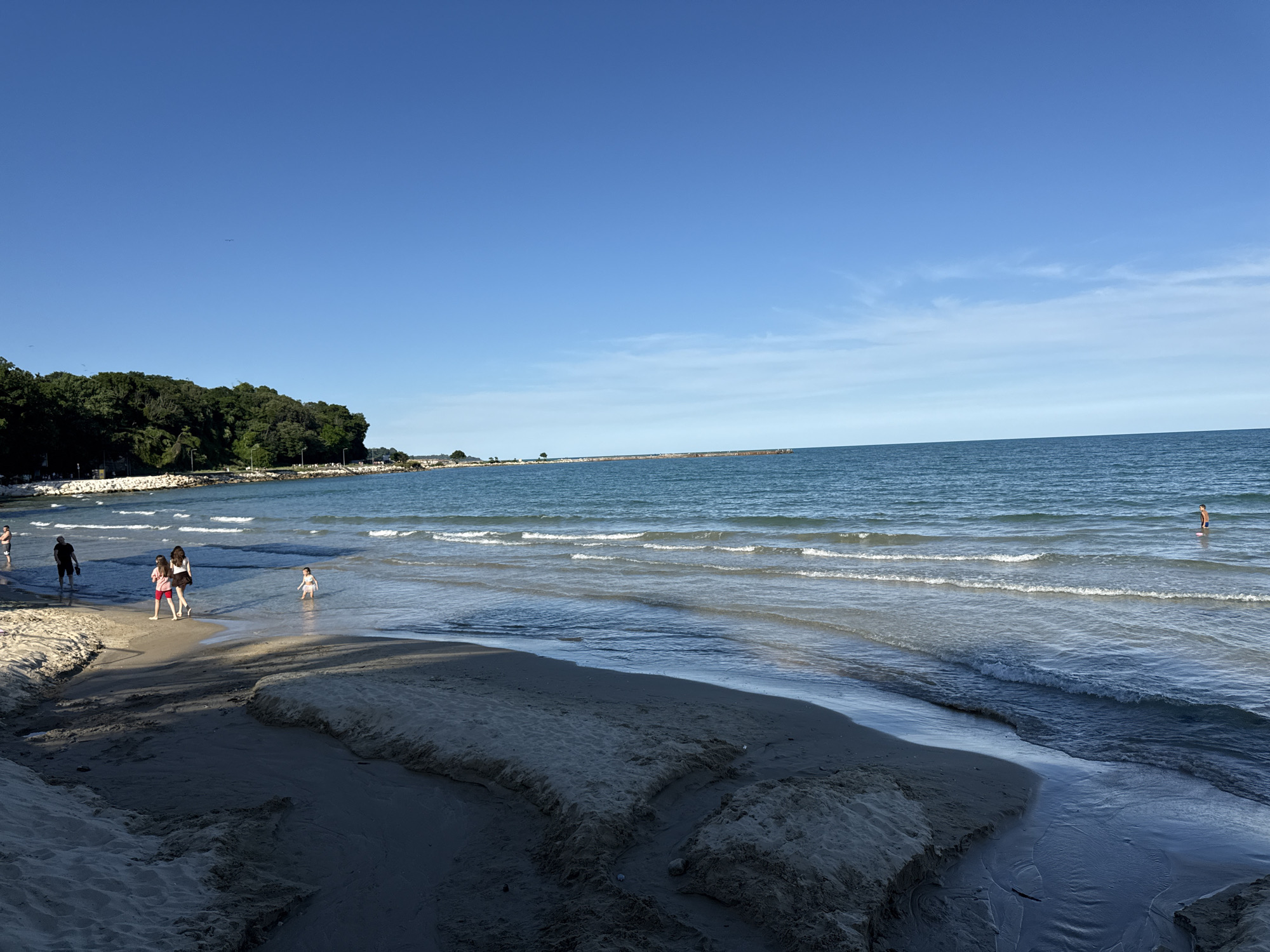 A group of people are enjoying a sunny day at the beach. The scene depicts a sandy shore with gentle waves lapping at the edge of the water. In the foreground, the wet sand shows some erosion, creating channels and mounds.
Several individuals are visible on the beach and in the shallow water. On the left, a man in a black t-shirt and shorts walks away from the camera. Further ahead, two young women, one in a white top and red shorts, and the other in a dark top and skirt, are walking towards the water. A small child in a white swimsuit is wading in the shallow water near them. On the right side of the frame, a single person stands in the deeper water.
The water is a deep blue, with white crests on the small waves. In the distance, a long pier or breakwater stretches out into the sea, and a line of trees and vegetation can be seen along the coastline behind the beach. The sky is a clear, vibrant blue with a few wispy clouds, suggesting a pleasant, sunny day. The lighting indicates it is likely late afternoon or early evening, with the sun casting shadows on the sand. There is no visible text in the image.
