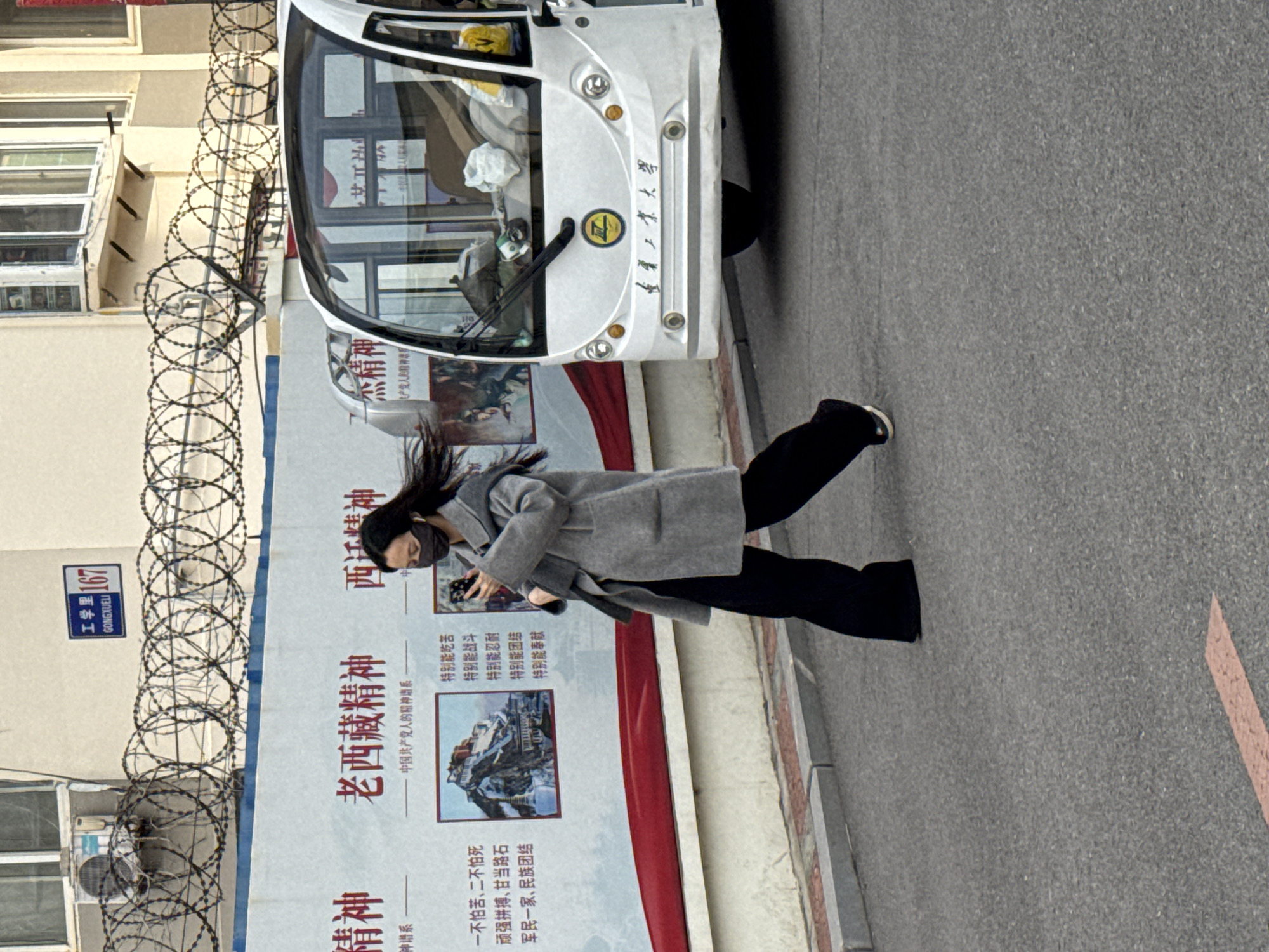 A street scene in what appears to be Zhongtun, China. A woman with long dark hair, wearing a gray coat and black pants, walks past a large poster on a building with razor wire. The poster has Chinese characters and images, possibly related to the "Old Tibet Spirit." A small white vehicle with Chinese writing on the front sits parked next to the poster. A street sign with "工学里 167 GONGXUELI" is visible on the building. The street and surrounding buildings appear to be urban.