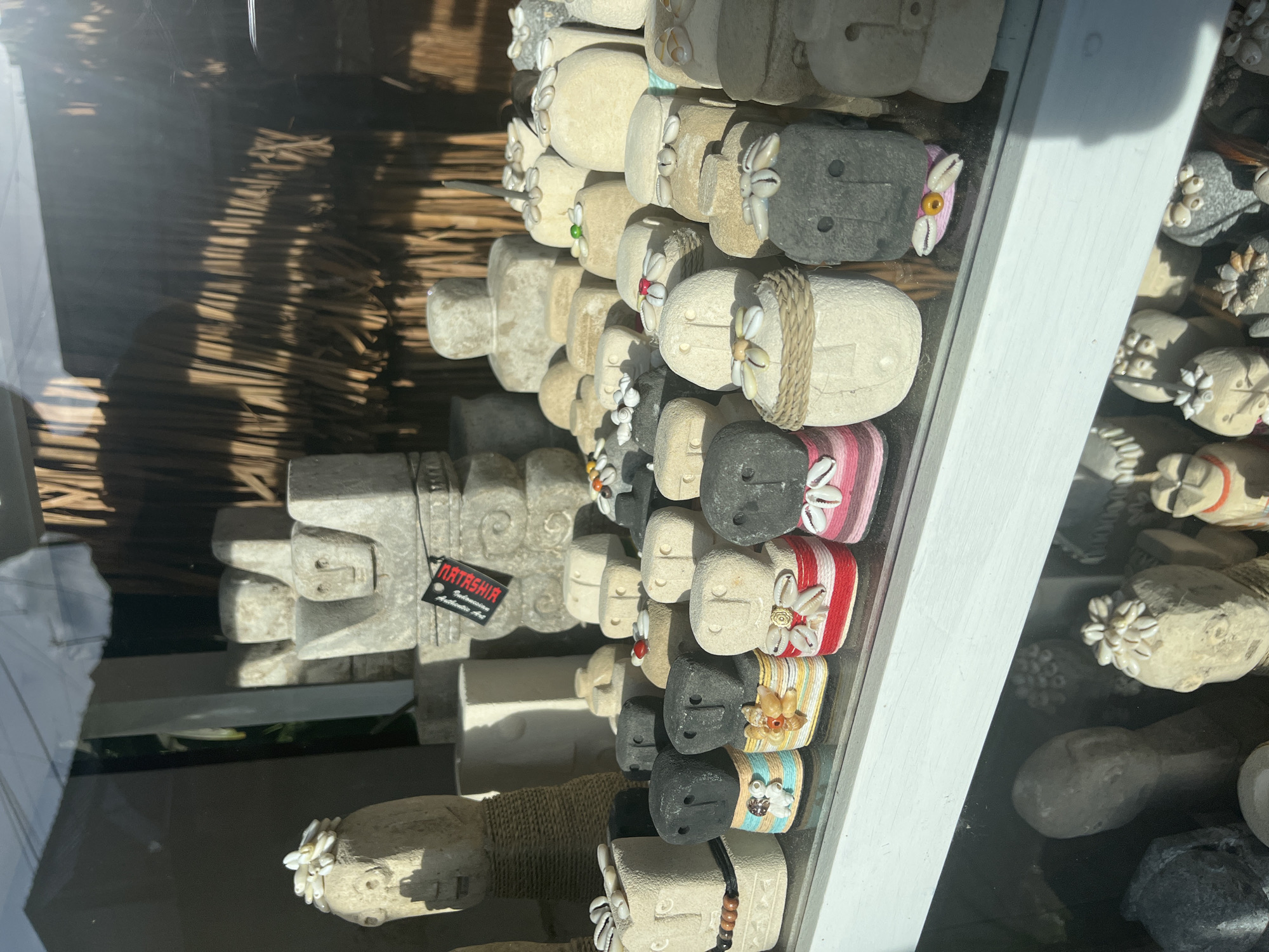 The image shows a display case in Ubud District, Indonesia, filled with small stone sculptures of heads. Some of the heads are decorated with shells, rope, or colorful fabric. A price tag is visible on one of the larger sculptures. The sculptures are displayed close together, and some are stacked on top of each other. A straw awning is visible in the background through the glass.