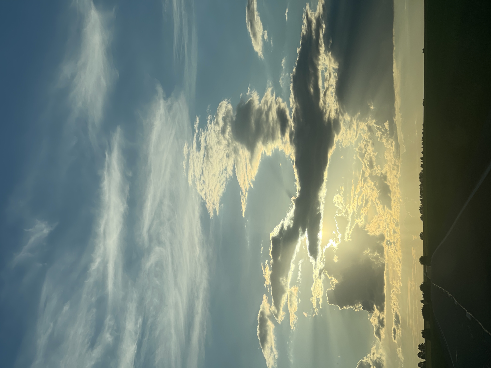 This media file displays a dramatic sky scene with a focus on clouds and sunlight. The image appears to be taken outdoors, likely in a rural or open area, as indicated by a dark, silhouetted strip of land along the right edge, which might be a field or a dark horizon.
The sky itself is a prominent subject. On the left side, wispy, white clouds are spread across a clear blue sky, giving a sense of a lighter, perhaps earlier or later part of the day. As the view moves to the right, the clouds become more dramatic and dense, with pockets of sunlight breaking through. These rays of light create a beautiful, almost ethereal effect, illuminating the edges of the darker clouds with a golden hue. The sun itself is mostly obscured by clouds, but its presence is strongly felt through the intense light.
The time of day is clearly suggested to be either sunrise or sunset due to the quality of the light and the way it interacts with the clouds, casting long rays and creating a warm, golden glow. The weather appears to be dynamic, with a mix of clear skies and significant cloud formations.
The dark strip on the right side of the image could be a treeline or the edge of cultivated land. A faint, light-colored line runs diagonally through this dark area, which could be a road or a path. The overall mood evoked by the image is one of awe and natural beauty, with the dramatic interplay of light and shadow in the sky. There is no visible text in the image.