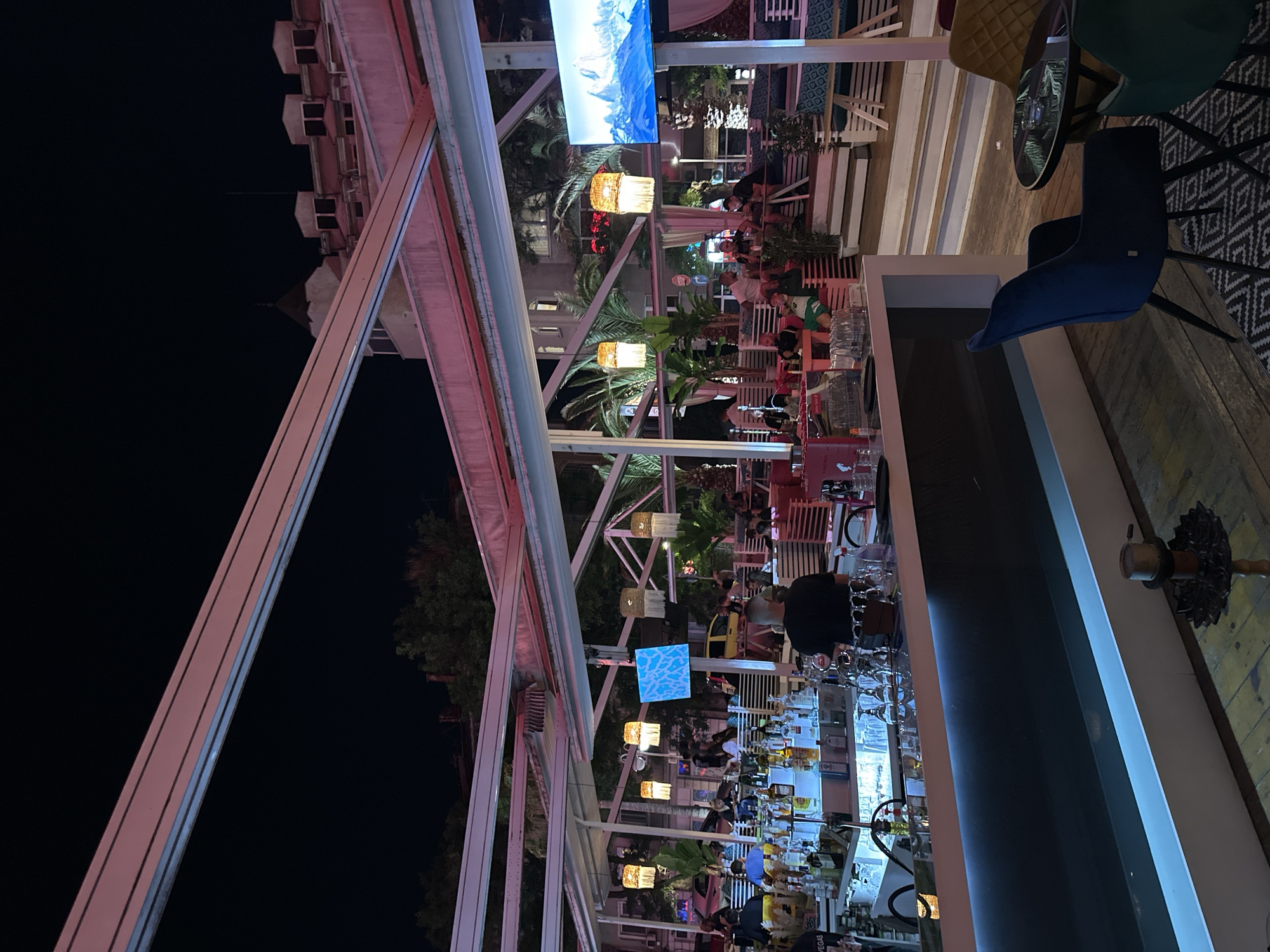 This image depicts a nighttime scene at an open-air bar or restaurant located in Plovdiv, Bulgaria. The establishment is covered by a retractable roof system, characterized by white structural beams illuminated with a pinkish hue. In the foreground, a sleek, modern bar counter with blue accent lighting showcases numerous liquor bottles, shakers, and glassware. A male bartender, wearing a dark shirt, is visible from behind, actively working at the bar.
Throughout the midground, patrons, primarily adults, are seated in various arrangements. These include tables with colorful chairs (blue velvet, yellow, green), and long, light-colored lounge benches. The seating areas are interspersed with decorative palm trees and other potted foliage. Overhead, multiple woven basket lamps emit a warm glow. Two large flat-screen televisions are mounted within the structure; the one on the left displays a blue, patterned texture, while the one on the right exhibits a scenic image of snow-capped mountains. The floor beneath the seating areas and bar is composed of wooden planks.
In the background, beyond the open-sided structure of the bar, urban elements are visible under a dark night sky. These include other buildings, streetlights, and the rear of a yellow taxi. A "STOP" sign is also partially visible on a pole. People within the bar are observed engaged in conversation and leisurely activities. On the wooden floor in the lower right, a hookah pipe is present.