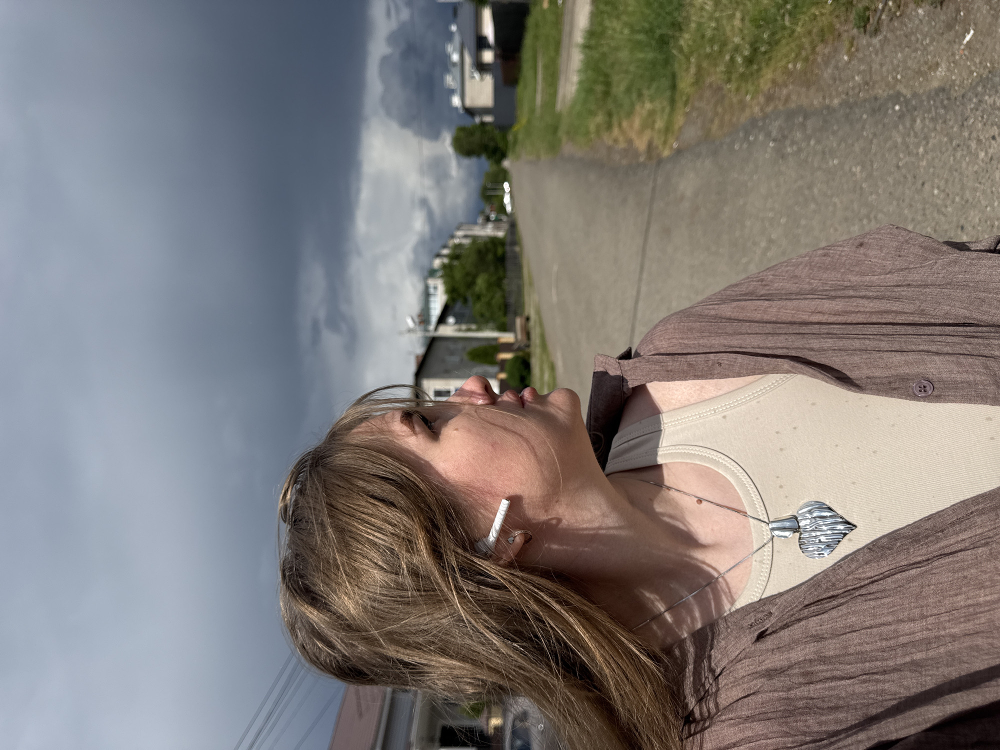 A medium outdoor shot depicts a woman, likely in her late 20s or early 30s, looking upwards. She wears a beige tank top, a brown button-down shirt, and a silver necklace with a distinctive pendant. She has long, light-brown hair, and what appears to be a white object in her left ear. The location seems to be a residential area with a sidewalk. Buildings are visible in the background, along with a cloudy sky. The perspective of the shot is unusual, as the camera is tilted. Given the street layout and architecture, the location Горный, Russia, is not conclusively identifiable.
