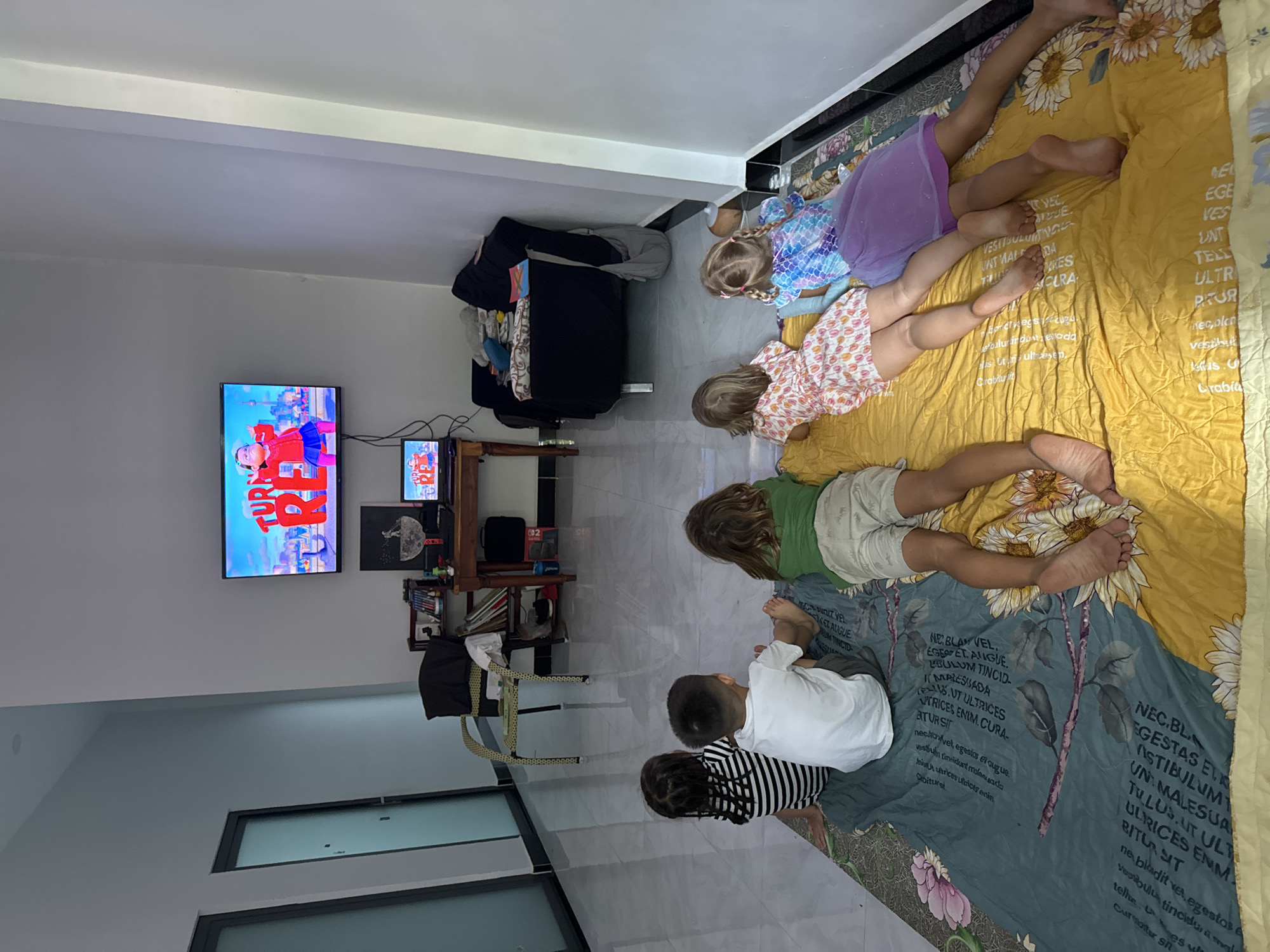 The image shows an indoor scene in what appears to be a living room, with several children lying on a blanket facing a television. The TV is mounted on the wall and displaying a cartoon. There are five children visible, ranging in age from toddler to pre-teen. They are wearing casual clothing, including dresses, shirts, and shorts. The blanket they are lying on has a sunflower pattern.
Behind the children, there's a TV stand with a smaller screen, some books, and other objects. A black armchair is situated against the back wall. To the left, there are windows and a door leading to another room. The overall color scheme of the room is light, with white walls and tiled floors.
While the location is specified as Mũi Né, Vietnam, there is nothing visually in the image that explicitly confirms this.