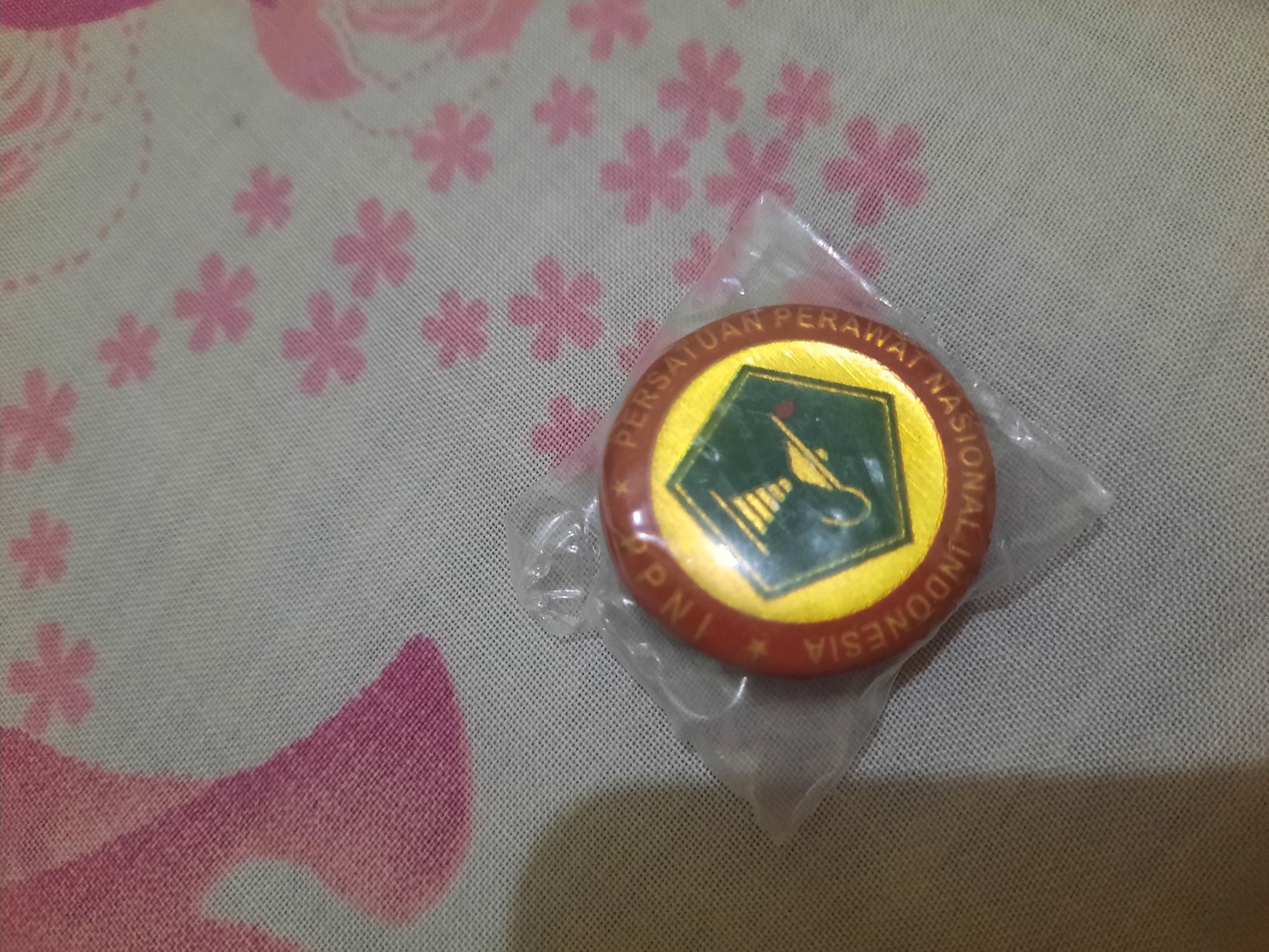 The image shows a round pin, partially in plastic wrapping, resting on a fabric surface. The pin has a red border with yellow text that reads "PERSATUAN PERAWAT NASIONAL INDONESIA" and "PNI". In the center of the pin is a green emblem with a stylized lamp. The fabric surface has a pattern of pink flowers and curved lines. The image is shot from a slightly overhead angle. There is no indication of the city of Lhokseumawe, Indonesia, in the provided image.