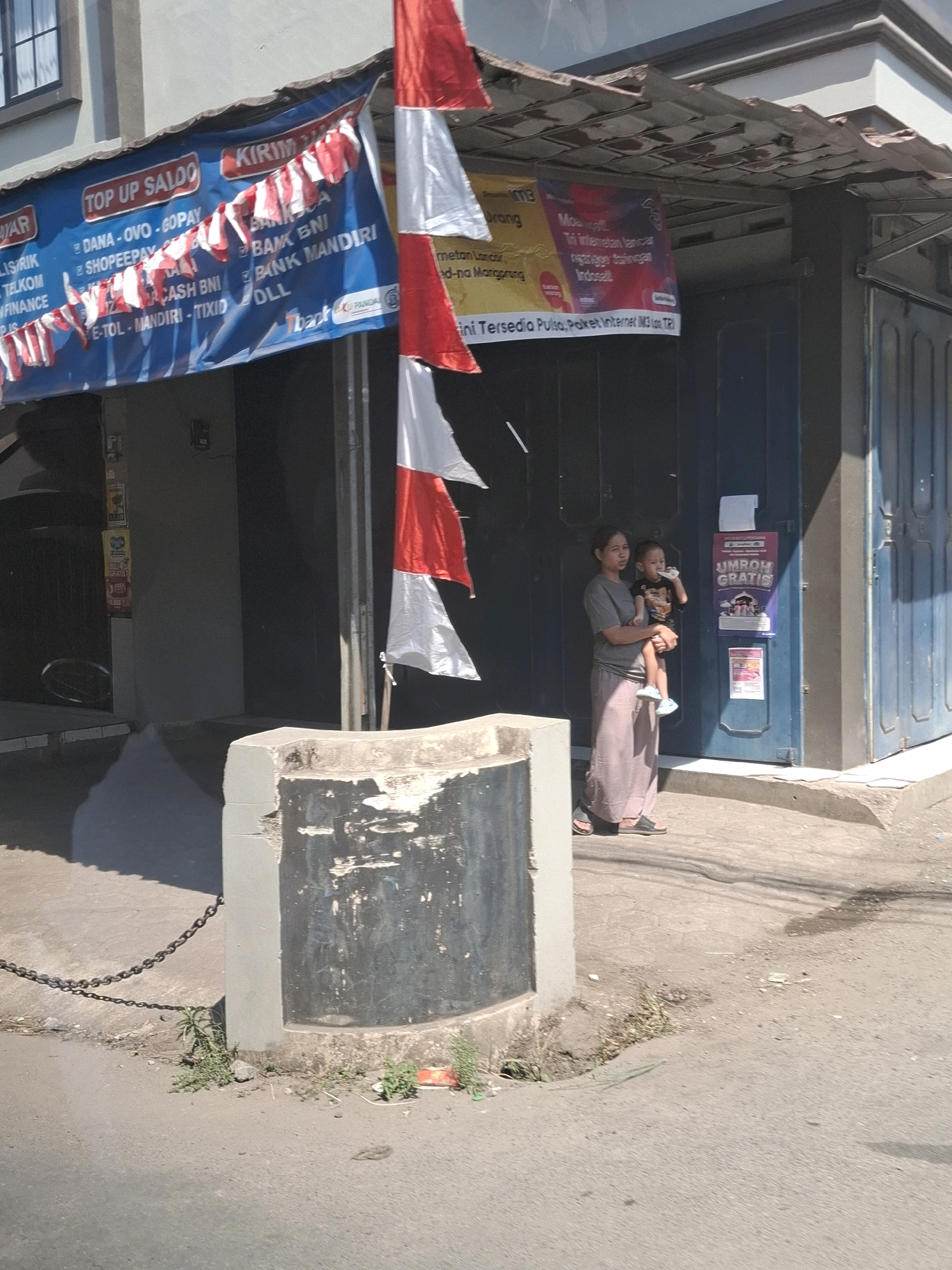This image depicts a street scene in Gunung Guruh, Indonesia. A woman is holding a young child, who is drinking from a bottle. They are standing on a paved sidewalk next to a building with a blue metal door. A red and white Indonesian flag is mounted on a pole nearby. In the foreground, a concrete structure with a metal chain is visible. A banner hangs from the building, advertising "TOP UP SALDO" and various payment methods. Another banner, partially visible, advertises "UMROH GRATIS" (Free Umrah). The ground is paved and shows some wear. The lighting suggests it is daytime, with shadows cast by the building and the flag.