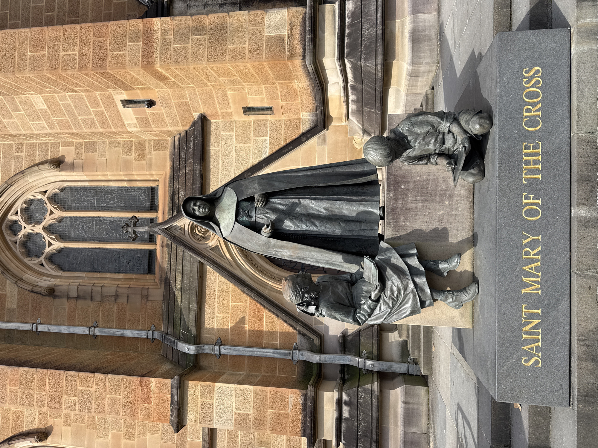 A bronze statue depicts Saint Mary of the Cross, a nun in a habit and wimple, standing with her hands clasped. At her feet are two children, also depicted in bronze. A young girl sits to the left, reading a book, while a young boy kneels to the right, writing on a slate. The base of the statue is inscribed with the words "SAINT MARY OF THE CROSS" in gold lettering. The statue is situated in front of a large, ornate building constructed of light-colored brick. The building features a large arched window with intricate tracery. The scene is set outdoors on a sunny day, casting shadows on the ground.