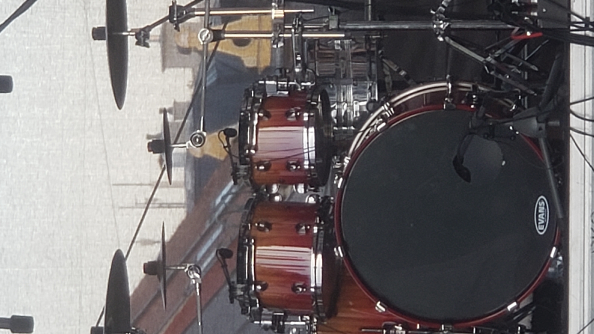 The image shows a drum kit on a stage. The drum kit includes a large bass drum with an "EVANS" logo, two tom-tom drums, and several cymbals. Microphones are attached to the tom-tom drums. In the background, there are buildings with red roofs and yellow accents, suggesting an outdoor setting, possibly a festival or concert. The overall lighting suggests it is daytime. The scene is devoid of people. This image was taken at the European Forum of New Films in Prešov, Slovakia, on September 22, 2023.