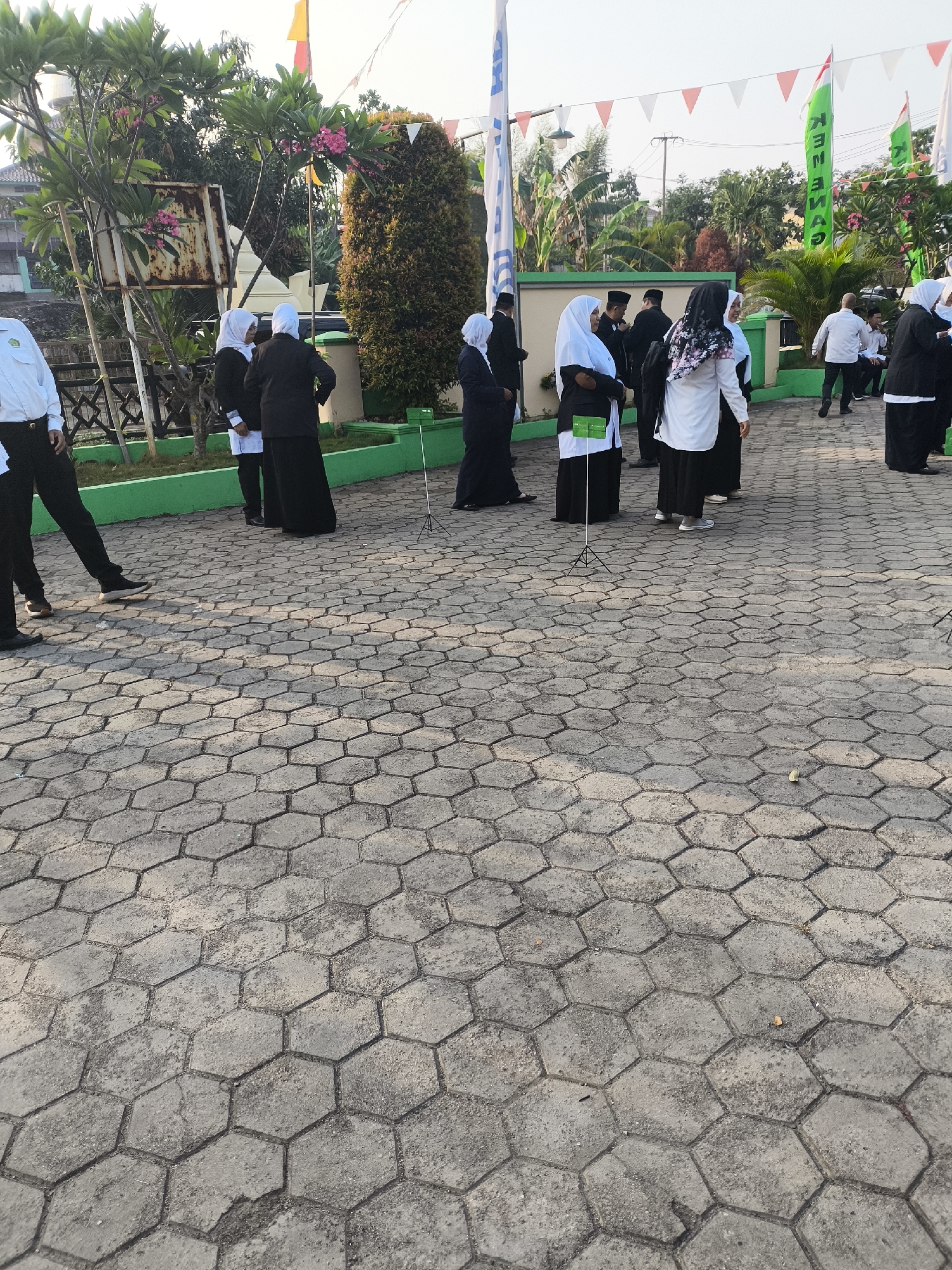 In Cilegon, Indonesia, a group of people, some wearing black and white uniforms and headscarves, stand on a hexagonal paver courtyard. The scene suggests an outdoor gathering or event. In the background, a low green wall separates the courtyard from a grassy area with trees and shrubs. Flags, some triangular and some vertical banners with green and white designs, are visible behind the people. The sky is bright, indicating it is daytime.