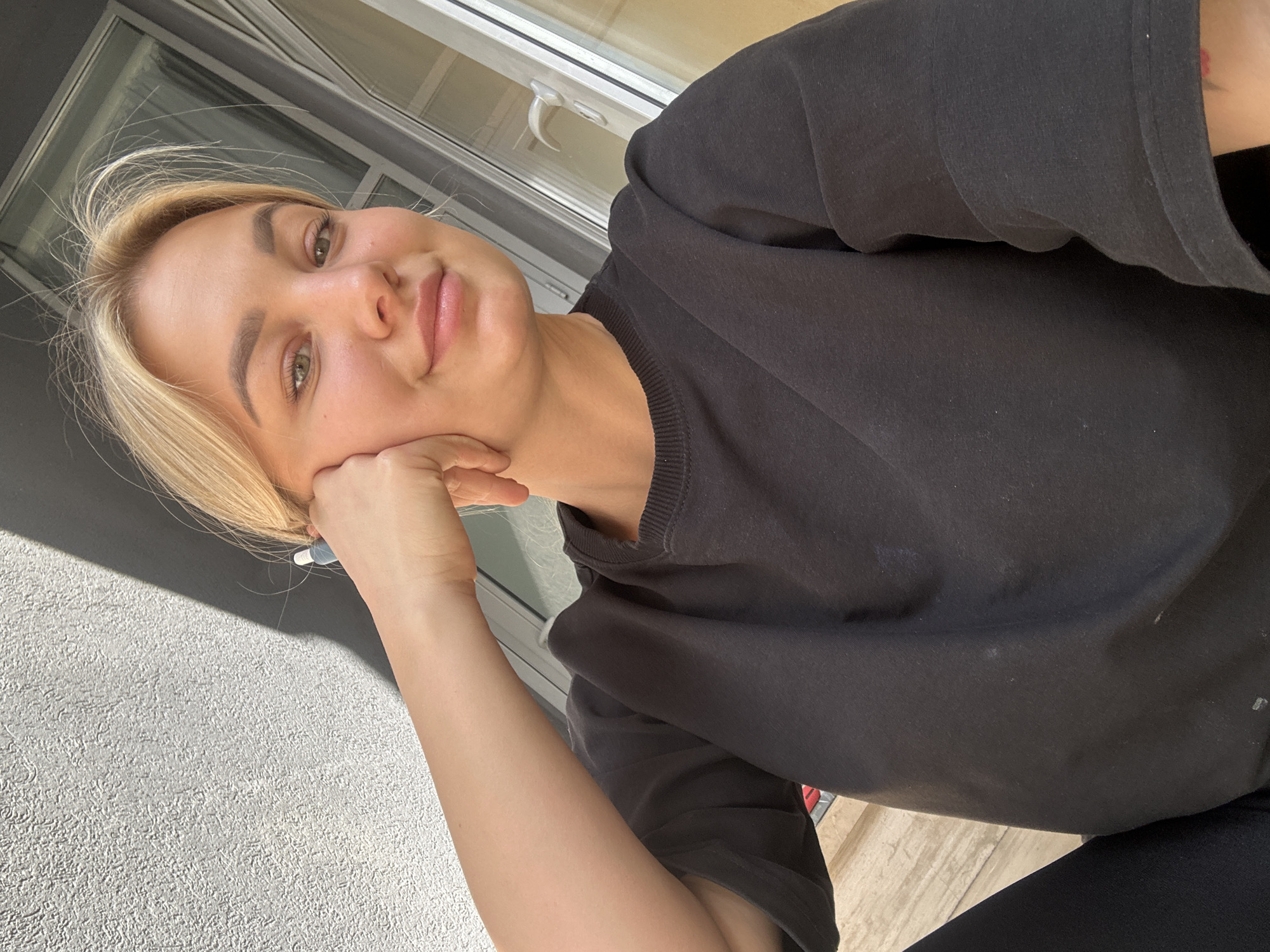A close-up, tilted portrait captures a young woman looking directly at the camera with a soft, relaxed smile. She has blonde hair, pulled back with some wisps framing her face, and light-colored eyes (greenish or hazel). Her left hand is propped under her chin, and a blue-tipped pen or stylus is tucked between her fingers.
She is wearing a loose-fitting, black crew-neck t-shirt or sweatshirt. The image is brightly lit by natural daylight, casting distinct shadows on a light-colored, textured wall to her left. In the background, a white window frame with a handle is visible, indicating an indoor setting, likely near a window. The bright light suggests a sunny day. The location is Budva, Montenegro. The woman appears calm and content. No visible text is present in the image.