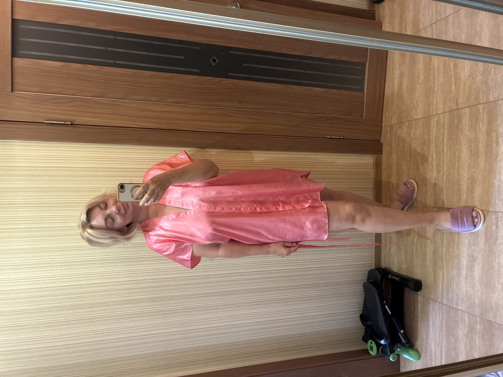 A woman stands in front of a mirror, taking a selfie. She is wearing a short, pink, satin robe and fluffy purple slippers. Her blonde hair is styled in a bob. The background consists of a textured wall and a wooden door. To her left, there is a compact exercise machine. The floor is tiled. This scene is likely indoors, possibly in a home or hotel room. There is no indication that this location is in Sochi, Russia.