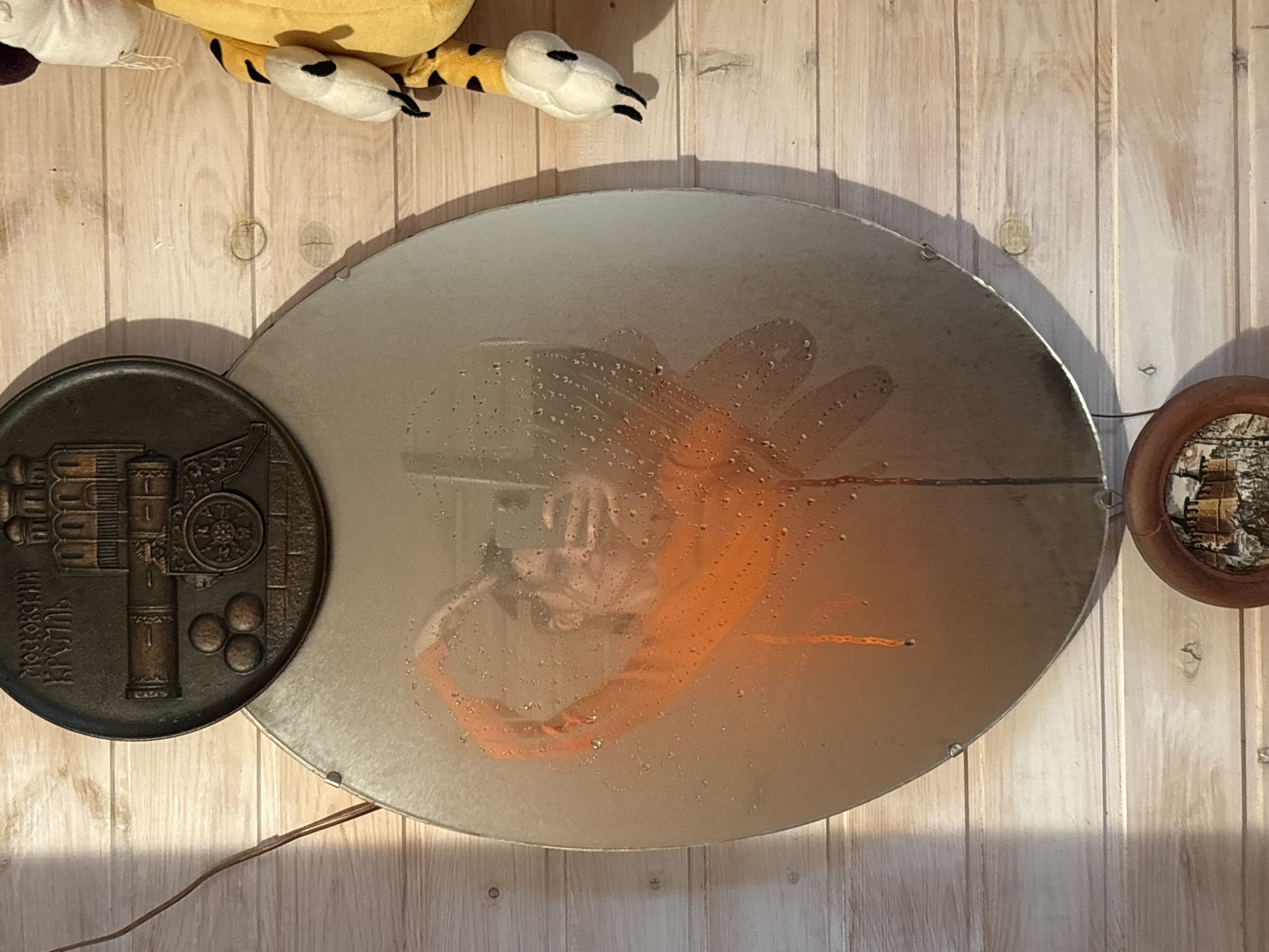 The image shows a still life with a few objects arranged on a light-colored wooden surface with vertical planks. In the upper left corner, there's a plush toy, possibly a tiger, with visible black stripes on its paw and a white foot. Below the toy, a decorative metal plate is positioned to the left. It is dark in color and appears to have a relief design of a building or machinery, along with Russian inscriptions. At the center of the frame is an oval-shaped object that seems to be a mirror or a reflective surface, partially covered with water droplets. The mirror is reflecting an orange-toned object, possibly clothing. To the right, another circular decorative plate or item with a wooden frame is visible. A thin, dark wire or cord runs along the lower left side of the frame, indicating an electrical component or connection. The location is Ленинградская, Russia.