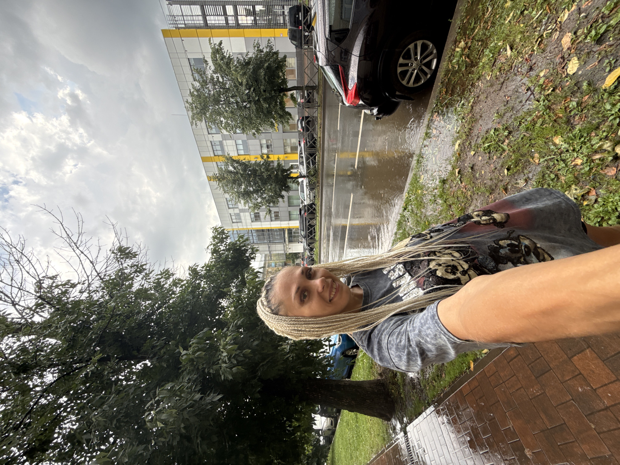 A woman with blonde braids is taking a selfie on a wet sidewalk in front of a modern building. The sidewalk is made of brick and is glistening with rain. To the right of the sidewalk is a grassy area with some fallen leaves, and beyond that, a street with several cars parked and driving. The street is also wet, with puddles reflecting the overcast sky. In the background, a multi-story building with white and yellow accents stands prominently. A large tree with green leaves is positioned to the left of the woman, partially obscuring the view of the building. The sky is cloudy, indicating a rainy or overcast day. The location is identified as Rusanovo, Russia.