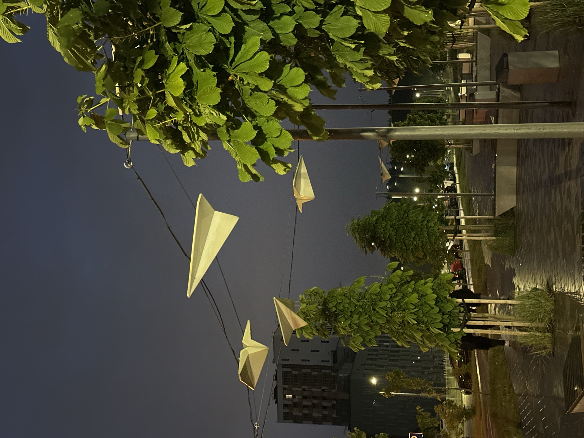 The image shows a view at night in Бараўлянскі сельскі Савет, Belarus. The view is mostly sky, with some buildings in the background. In the foreground are some trees and streetlamps with paper airplane decorations. The streetlamps are spaced out along a paved walkway, and there are some benches along the walkway. The light is dim, but the streetlamps provide some illumination. There are no people visible in the image.