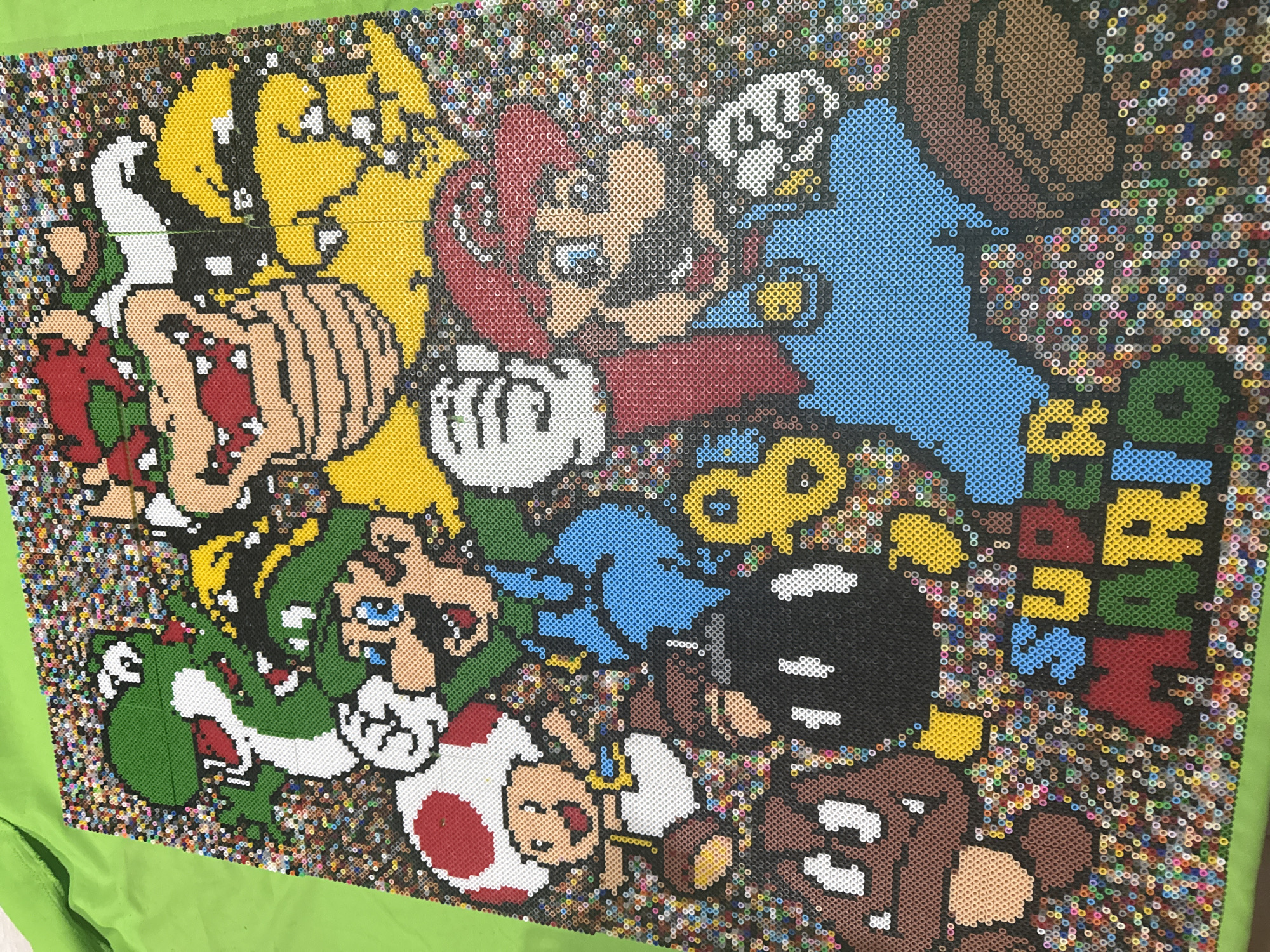 The image depicts a pixelated representation of Super Mario World characters and logo, constructed from small, circular beads. Characters include Mario, Luigi, Yoshi, Toad, Bowser, Goomba, and a Bob-omb. The backdrop consists of a multitude of similarly sized beads in various colors, creating a speckled appearance. The artwork is placed on a green fabric surface. There is no indication of the image location being Puebla, Mexico.