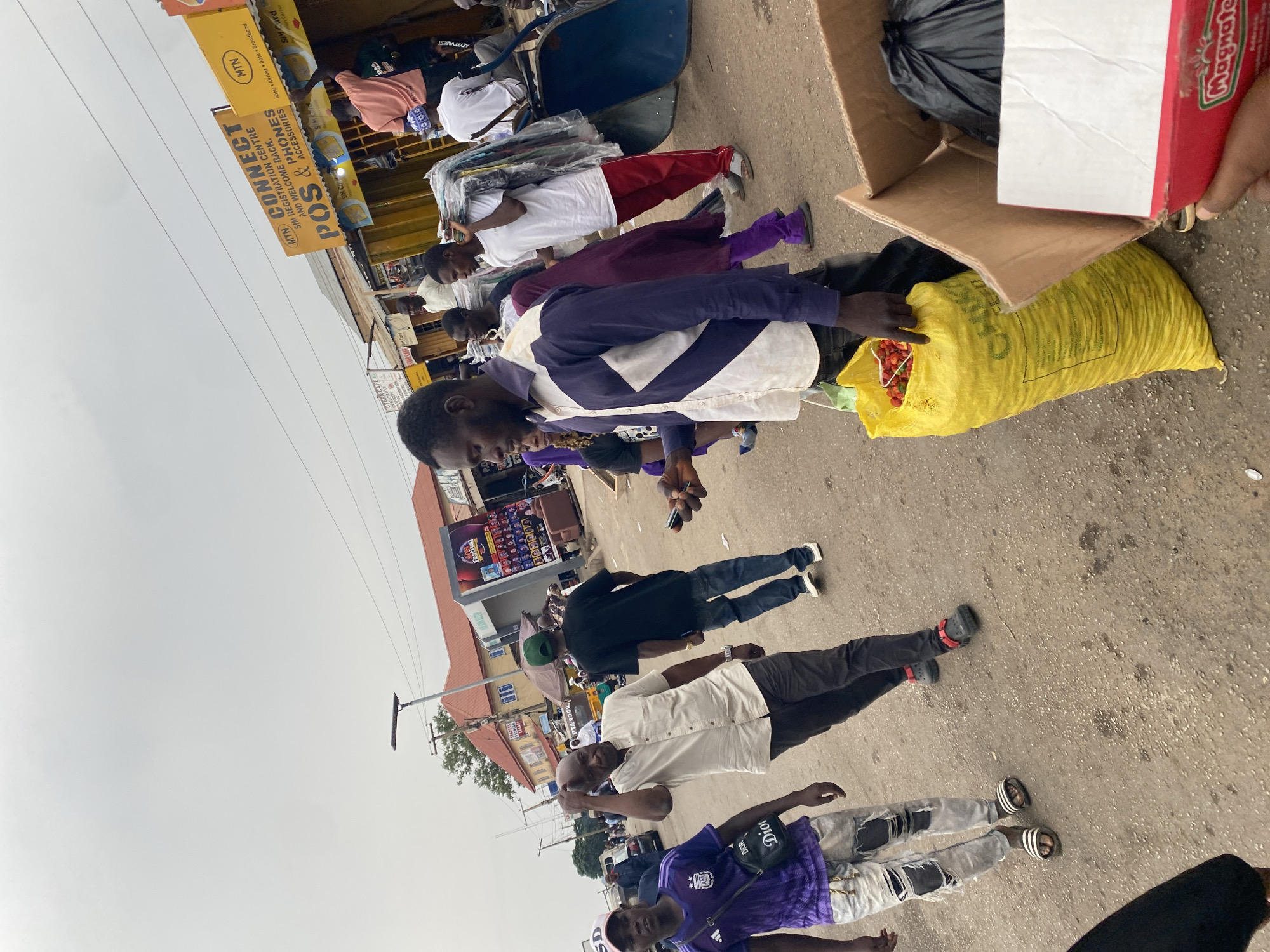 The image depicts a street scene in Gura Top, Nigeria. Several individuals are visible, some carrying goods or engaged in commerce. One person appears to be transferring items from a yellow sack into a cardboard box. The background shows street vendors, signage for "MTN CONNECT" (a SIM registration center), and a "Cyber Cafe" sign. A "Magnate" branded box is also visible. The environment is an urban street setting with signs of commerce and pedestrian activity.