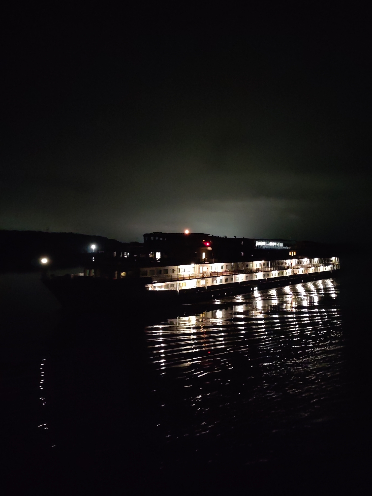 A riverboat, illuminated with interior lights, is docked on a dark body of water. The reflection of the boat lights ripple on the water's surface. A dark, slightly visible shoreline is in the background. It is nighttime. The location is the city of Unknown, Unknown.