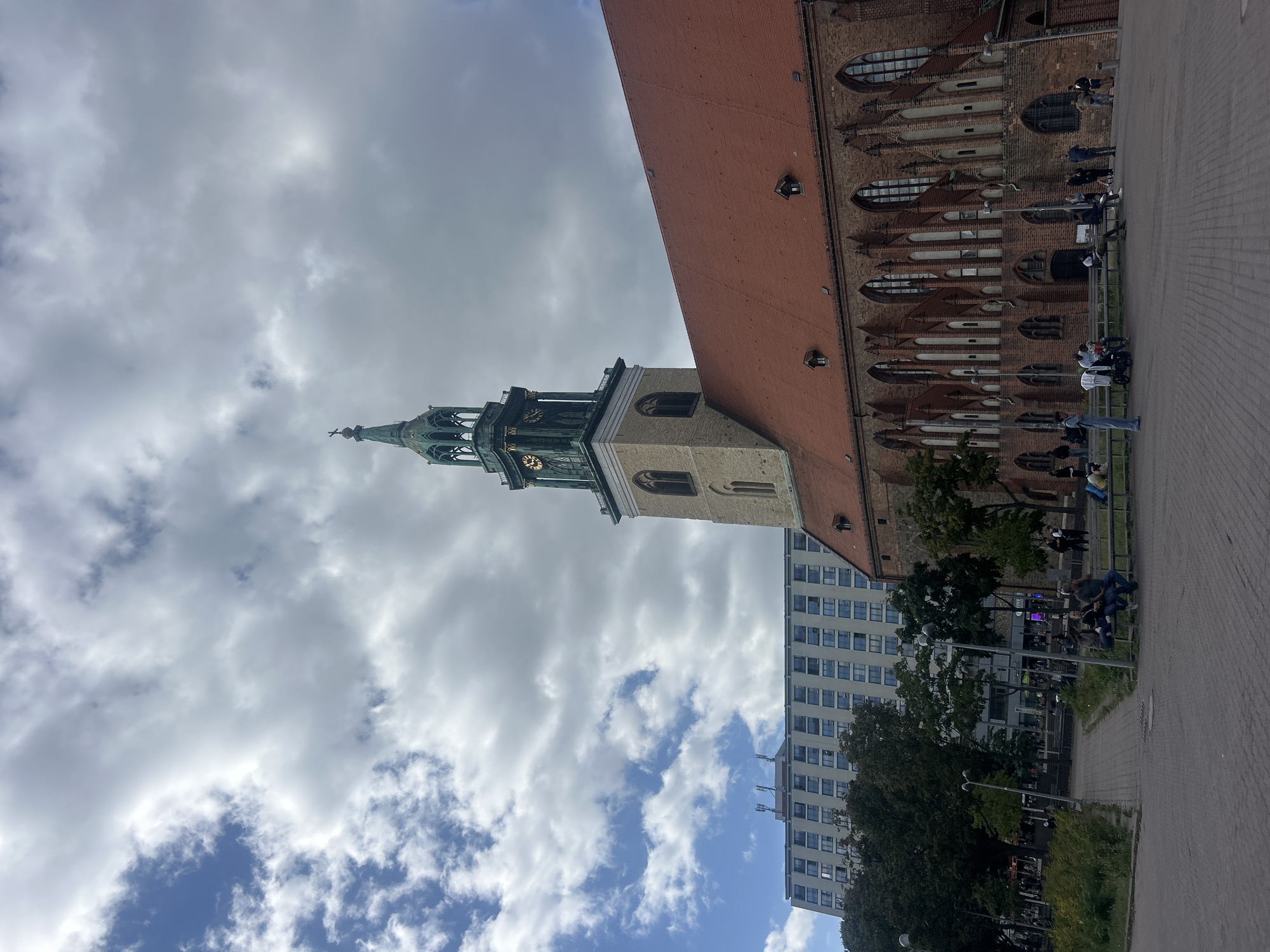 The image depicts a view of the Marienkirche (St. Mary's Church) in Berlin, Germany, with a modern high-rise building visible in the background. The Marienkirche is a prominent brick Gothic church with a tall, green-spired clock tower. Its red tiled roof is visible, along with the arched windows and buttresses of its facade. In front of the church, a paved public area is populated by several individuals sitting on benches and standing around. Trees and streetlights are also present. The sky is cloudy, with patches of blue visible. The modern building behind the church is a multi-story structure with numerous windows.