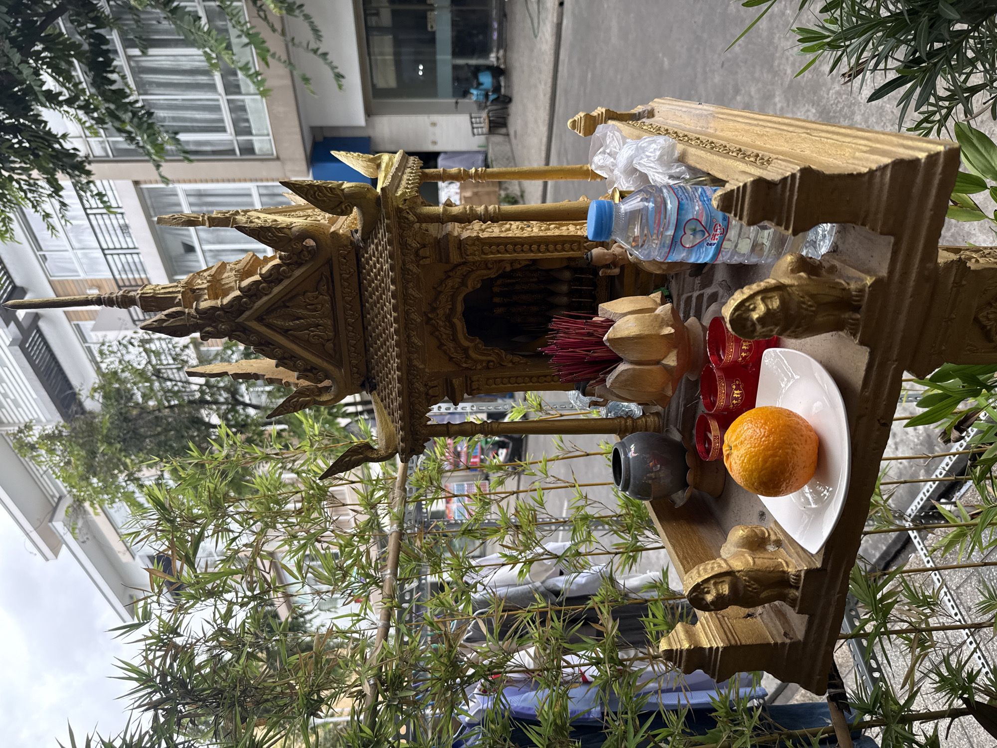 A golden Buddhist altar is situated on a street in Khan Chbar Ampov, Cambodia. The altar features decorative elements, including what appear to be lion statues on either side. On the altar, there is an orange on a white plate, several small red cups, a bundle of incense sticks, a small figurine of a person, and a bottle of water. Behind the altar, bamboo plants are visible, along with buildings and a blue scooter in the background.