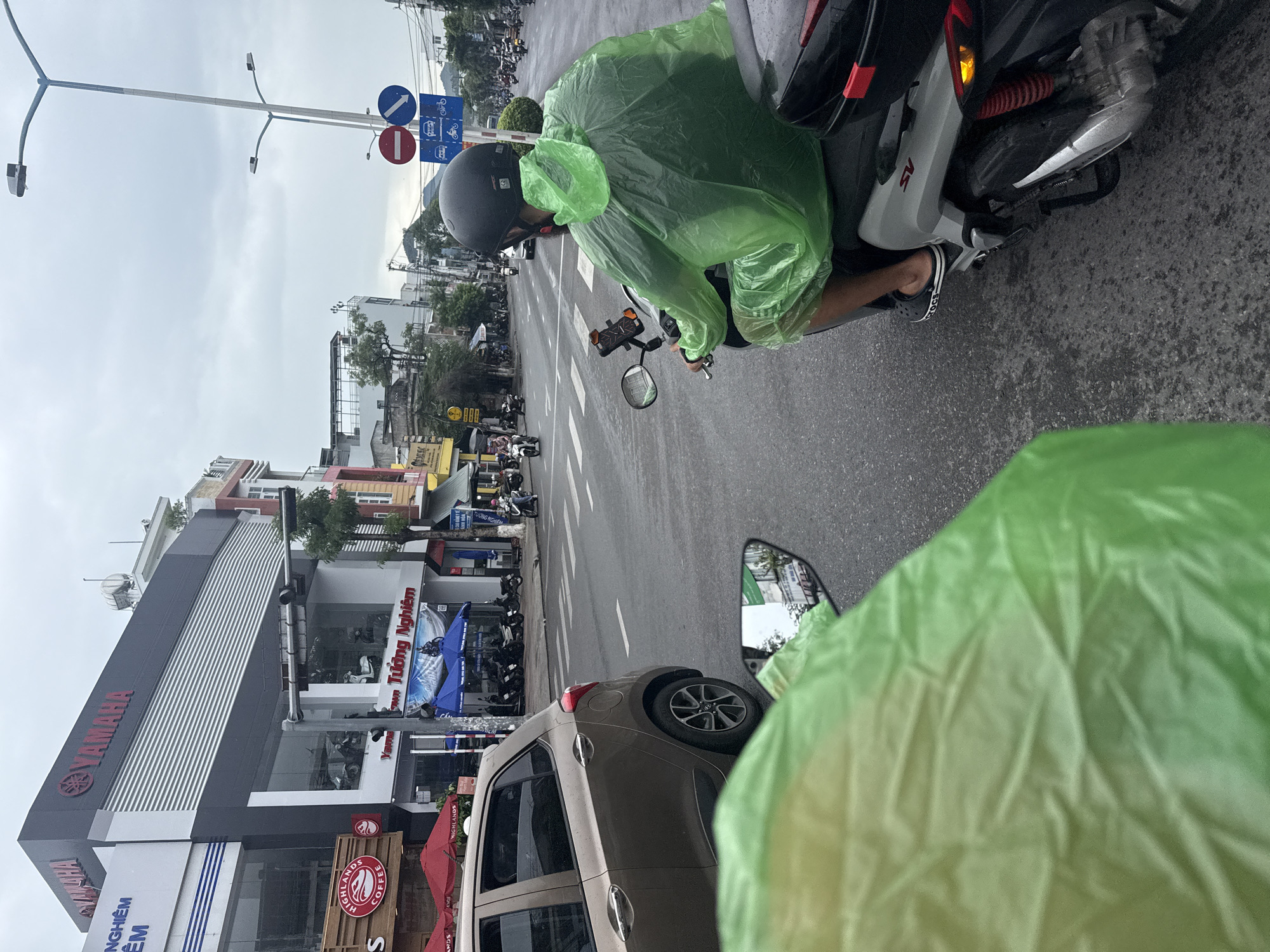 The image depicts a street scene in Nha Trang, Vietnam, likely taken on a cloudy or overcast day. In the foreground, a person wearing a bright green rain poncho is seated on a scooter, viewed from behind and slightly to the side. The scooter is silver with visible "SV" lettering and red suspension springs. The person is wearing dark pants and black Crocs sandals. A second person, also in a green poncho, is visible in the immediate foreground, partially obscuring the view and appearing to be on a second scooter, suggesting the photo was taken from a moving vehicle.
The middle ground shows a street with a pedestrian crossing, a beige car parked on the left, and several other scooters and motorcycles parked or in motion. On the right side of the street, a building with a prominent "YAMAHA" sign and logo is visible, indicating a Yamaha dealership or service center. Below the Yamaha sign, the text "TƯỜNG NGHIÊM" is displayed, likely the name of the dealership. Red umbrellas are set up outside a "HIGHLANDS COFFEE" establishment to the left of the Yamaha building.
Further down the street, other buildings, businesses, and traffic can be seen. Traffic signals and street lights are present, with a sign showing prohibited and permitted directions of travel for different vehicle types. The overall environment suggests a typical urban street with commercial establishments and active traffic.