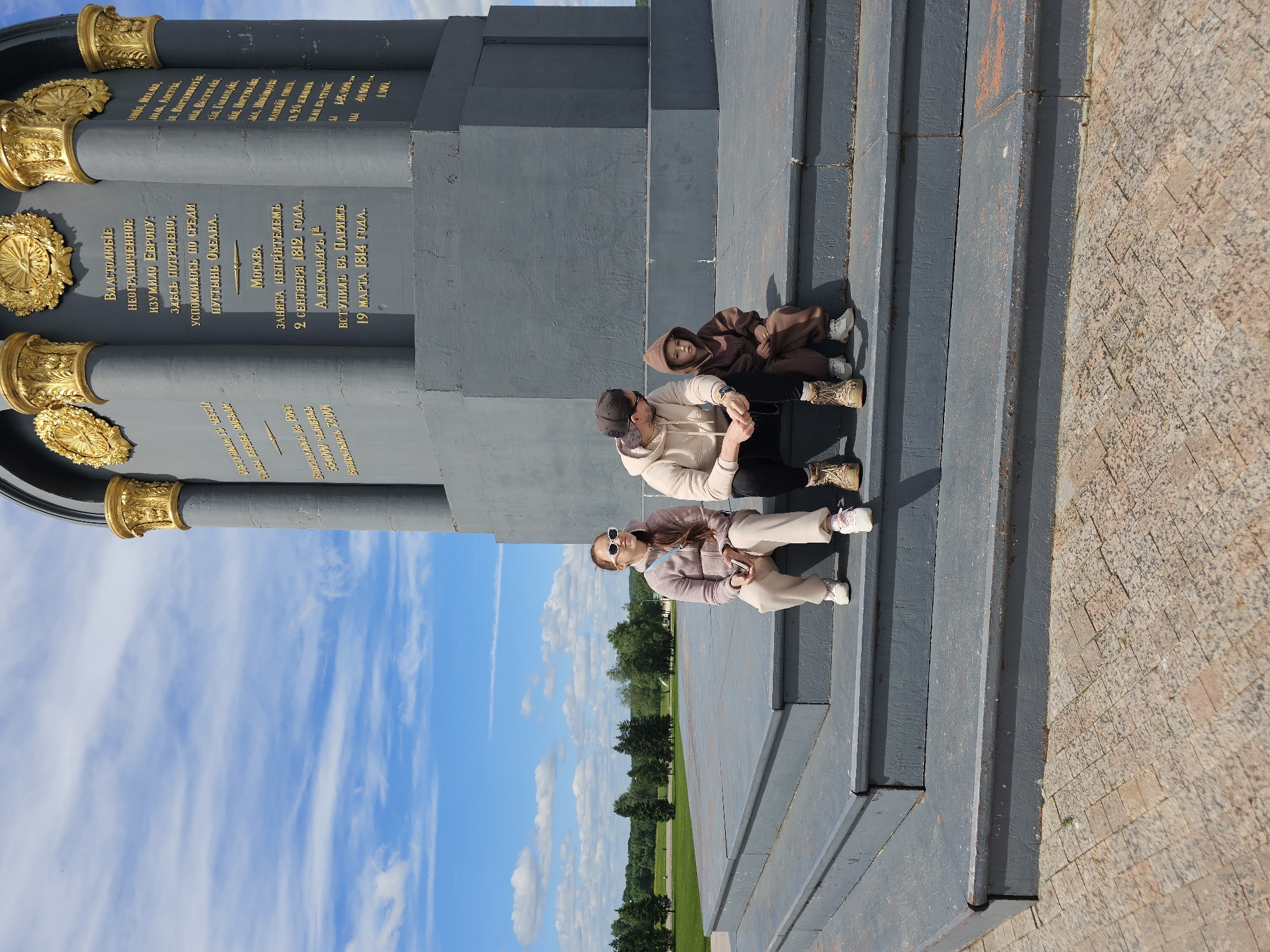 Here is a description of the image:
The image depicts three people sitting on the steps of a monument on a sunny day. The monument features gray columns with golden ornaments and inscriptions in Russian. The people are a woman wearing sunglasses and a pink outfit, a man in a beige hoodie and black pants, and a child in a brown hoodie. They are sitting on the gray steps leading up to the monument. In the background is a view of trees and a blue sky with clouds.