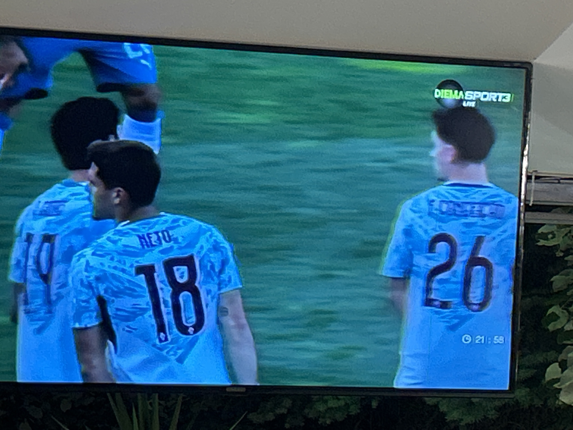 A flat-panel television screen, displaying a live soccer match on a green field, dominates the frame. Four male players are visible from behind, all wearing light blue jerseys with a subtle patterned design. In the central foreground, a player with dark hair has "NETO" and the number "18" clearly visible on his back. To his immediate left, another player is partially seen, wearing a jersey with the number "10" visible. Another player's dark hair and back are dimly visible behind the "NETO 18" player, further to the left. On the right side of the screen, a player with dark hair wears jersey number "26" and the name "LOBELL" printed above it. In the upper right corner of the TV screen, the text 'DIEMASPORT3 LIVE' is displayed, identifying the broadcast channel. A small clock icon and the time '21:58' are visible in the bottom right corner of the screen. The television is situated in what appears to be a covered area, possibly indoors or on a balcony, with a dark border at the bottom. Foliage, consistent with plants or tree leaves, is visible along the right side of the physical frame, partially obscuring the background behind the screen. While the broadcast channel, Diema Sport 3, is a Bulgarian network, the image contains no specific visual cues or landmarks to identify a precise location within Blagoevgrad, Bulgaria.