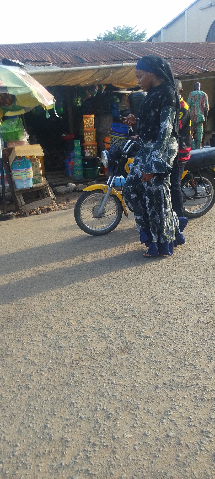The image, likely taken in Lokoja, Nigeria, shows a woman standing next to a yellow motorcycle on a paved road. She is wearing a long, patterned dress and a head covering. A man is also visible on the motorcycle, wearing a black shirt. Behind them is a storefront with various containers and goods displayed. The environment suggests an outdoor market or commercial area. Two additional men, wearing traditional clothing, are near the storefront. The lighting indicates it is daytime, with shadows cast across the road. The road surface appears rough and textured.