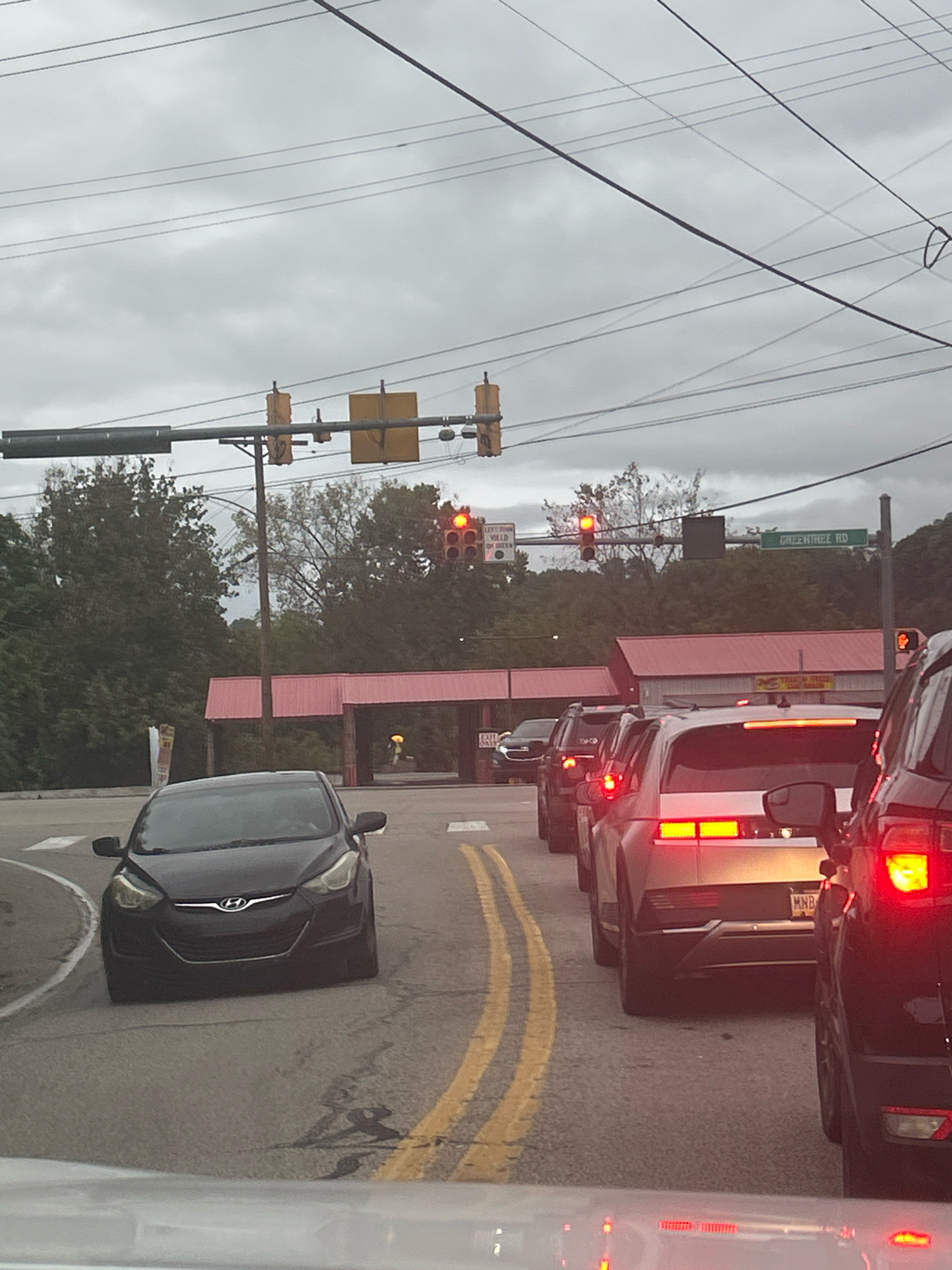 A traffic jam is depicted at an intersection in Scott Township, Pennsylvania, USA, under an overcast sky. Several cars are stopped in multiple lanes on a wet asphalt road marked with yellow double lines. In the background, a building with a red roof is visible, and trees line the horizon. Traffic lights are illuminated red, indicating that traffic is stopped. A green street sign reads "GREENTREE RD". The composition suggests the viewer is in a vehicle, observing the traffic ahead.