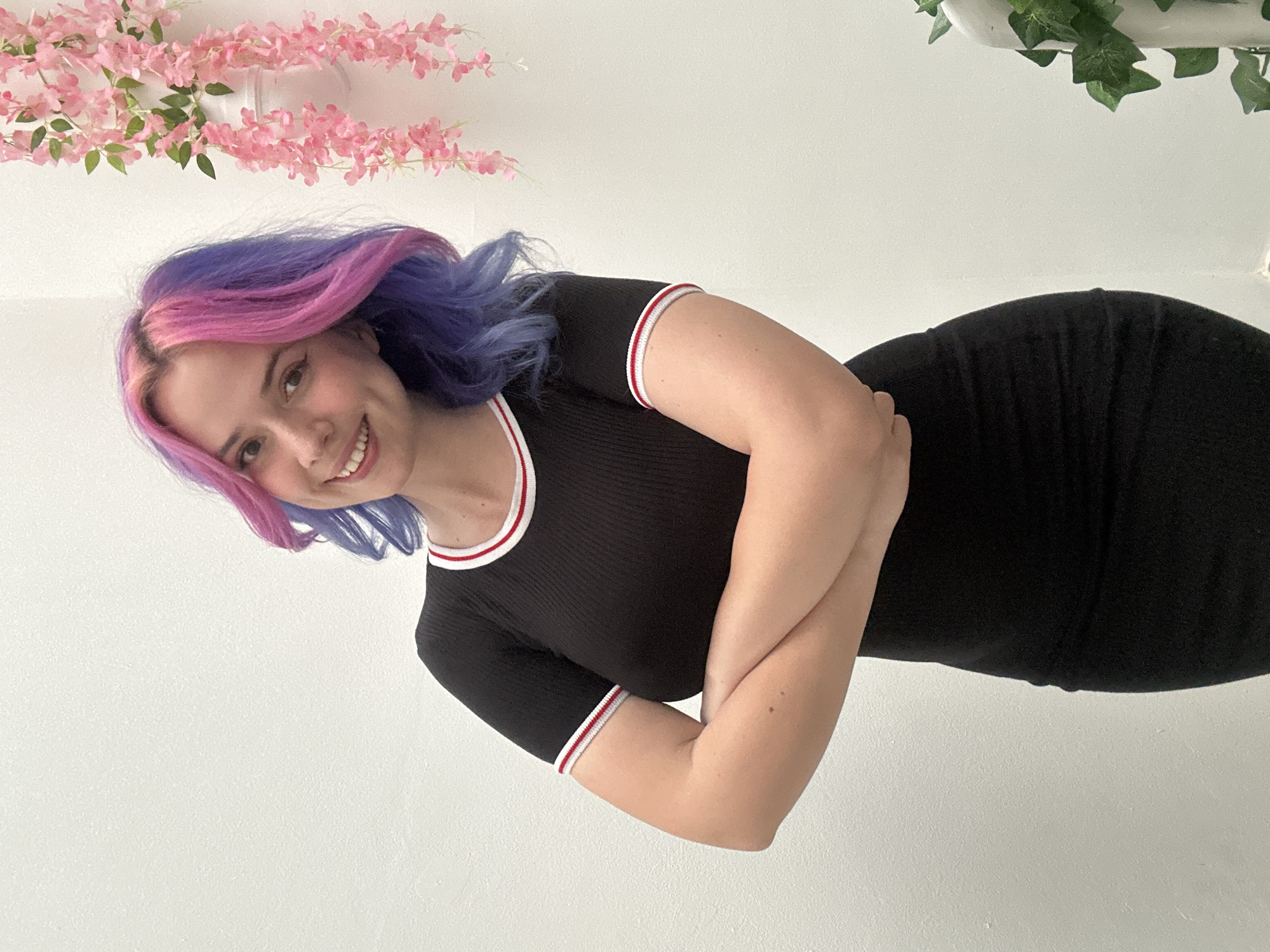 A young woman with distinctive two-toned hair, featuring bright pink on one side and blue-purple on the other, is captured in a half-body shot. She is smiling broadly, showing her teeth, and her expression conveys happiness and friendliness. Her arms are crossed comfortably in front of her body. She is wearing a form-fitting, black ribbed short-sleeved top or dress with white and red striped trim along the collar and the sleeve cuffs.
The background is a plain white wall, suggesting an indoor setting. In the upper left part of the frame, a cluster of artificial pink floral garlands, possibly suspended from above, adds a decorative element. On the upper right, artificial green ivy foliage is also visible, contributing to the indoor decor. The lighting is even and typical of an interior space, offering no specific clues about the time of day or external weather. There is no visible text in the image.