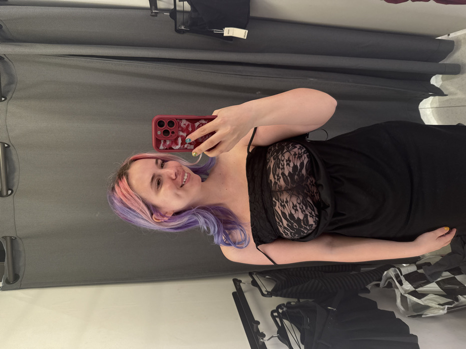 A female individual is depicted in a mirror selfie taken within what appears to be a retail changing room. The specific location within the City of Zagreb, Croatia, cannot be determined from the provided image.
**Person:**
The subject is a light-skinned female, smiling at her reflection. She has long, wavy hair, dyed with pink streaks at the front/top blending into purple. Her visible fingernails on her left hand are painted yellow. She is wearing a black, off-the-shoulder style dress, which is pulled down to reveal a black lace garment underneath. This lace garment is transparent across her chest. The thin straps of the black dress hang off her shoulders. She holds a red smartphone with a distinct red and white patterned case featuring a multi-lens camera array in her left hand, angling it to capture the selfie.
**Environment and Objects:**
The background features grey fabric curtains, suspended from a black rod with rings, typical of a fitting room. To the left, a black metal clothing rack is visible, holding various dark-colored garments (skirts and tops). A black and white patterned shopping bag rests on the floor beneath the rack. To the far left, a plain white wall is visible. To the right, another section of a black clothing rack is partially visible, displaying a black garment with a white tag attached. The floor is light-colored.