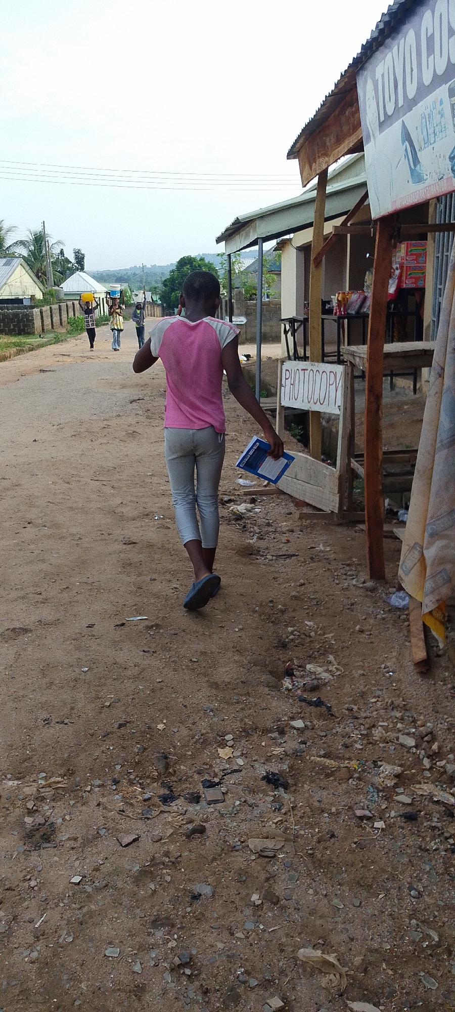 The image captures a street scene in Adankolo, Nigeria. A young person walks away from the viewer, holding a blue folder in their left hand. They wear a pink and white shirt, grey trousers, and blue sandals. In the background, three people walk in the same direction, carrying items on their heads. To the right, there is a small storefront with a sign that reads "PHOTOCOPY." A sign above the storefront says "TOYO CO." The ground is dirt with some debris scattered around. Buildings and trees are visible in the background, under a somewhat overcast sky.
