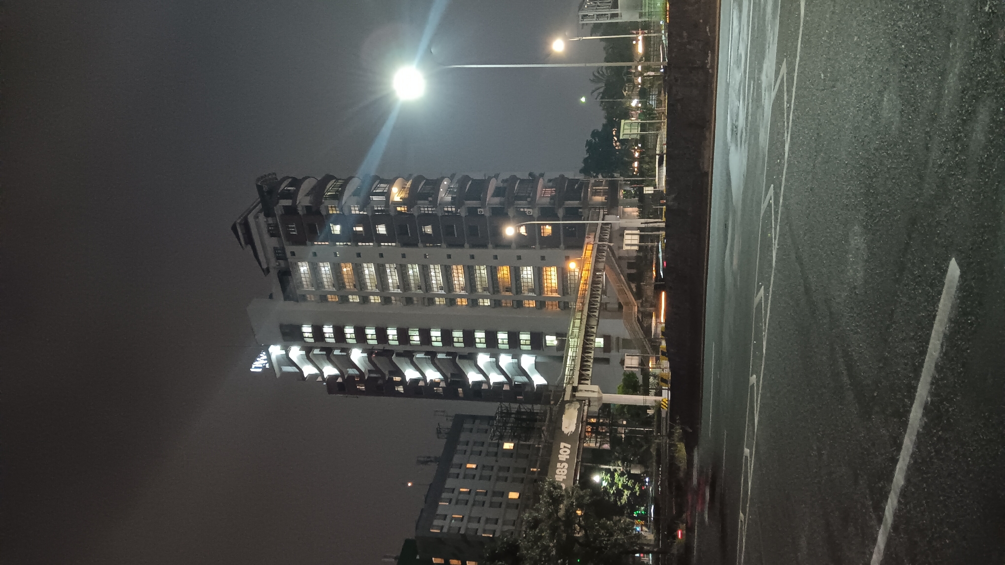 The image shows a modern apartment building at night, identified as being located in South Jakarta, Indonesia, specifically along Jalan Casablanca Raya. The building is tall and illuminated by numerous windows emitting a warm, yellowish light. Its facade features a distinctive tiered design with balconies and cantilevered sections. A prominent sign with white, illuminated lettering, appearing to read "TW...", is visible at the top of the building.
In the foreground, a multi-lane asphalt road is visible, with white lane markings and some puddles, suggesting recent rain or damp conditions. The road appears to be a major thoroughfare, as indicated by the presence of streetlights and some blurry streaks of light that suggest moving vehicles.
Above the road, an elevated pedestrian walkway or bridge is visible, connecting to the apartment building. This walkway is also illuminated, with stairs leading up to it. Next to the walkway, a dark-colored structure with the numbers "485 407" stenciled on it is present, possibly a part of the infrastructure or signage for a transportation system.
To the left of the main apartment building, another, slightly shorter building with a similar pattern of illuminated windows is visible, suggesting a dense urban environment. Trees and other vegetation can be seen around the base of the buildings and along the roadside. The overall atmosphere is that of a busy urban area at night, characterized by artificial lighting against a dark, possibly overcast, sky.