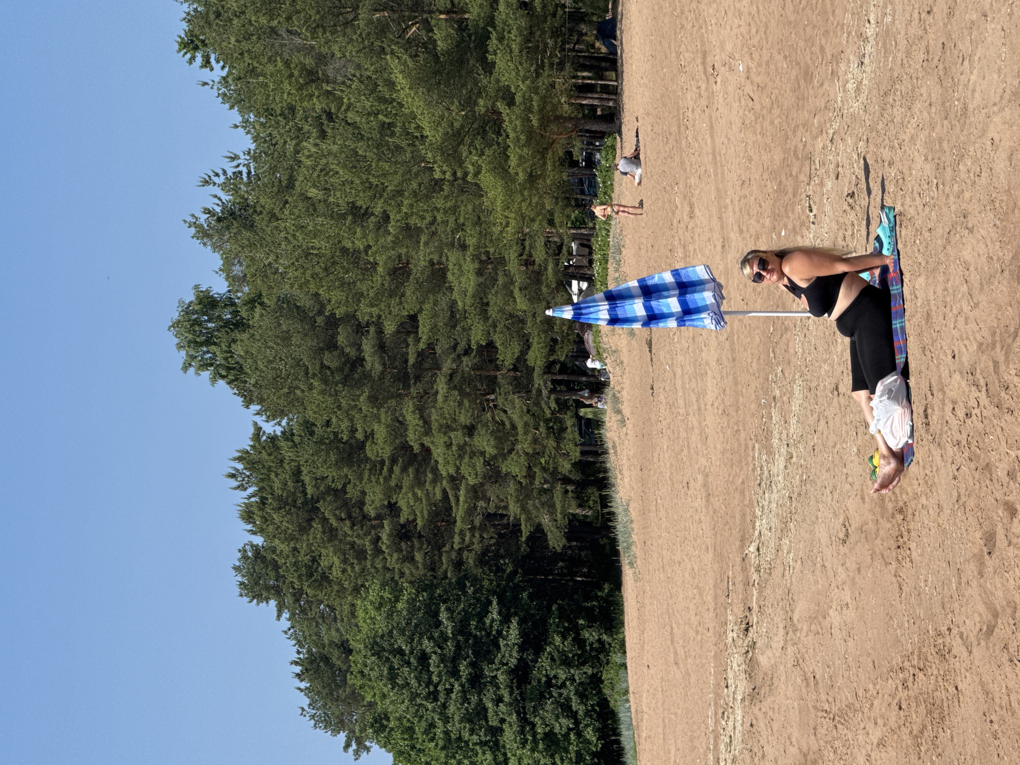 A woman sits on a beach in Zelenaya Roshcha, Russia, on a sunny day. She is positioned on a multicolored mat on the sand, leaning back slightly with her legs extended. She is wearing a black sports bra and black leggings, with sunglasses on. A blue and white striped beach umbrella is positioned behind her. Nearby, her light blue Crocs are placed on the sand. In the background, a dense forest of green pine trees forms a backdrop to the sandy beach. The sky is clear and blue. Other beachgoers are visible in the distance, with some tents and cars parked amongst the trees.