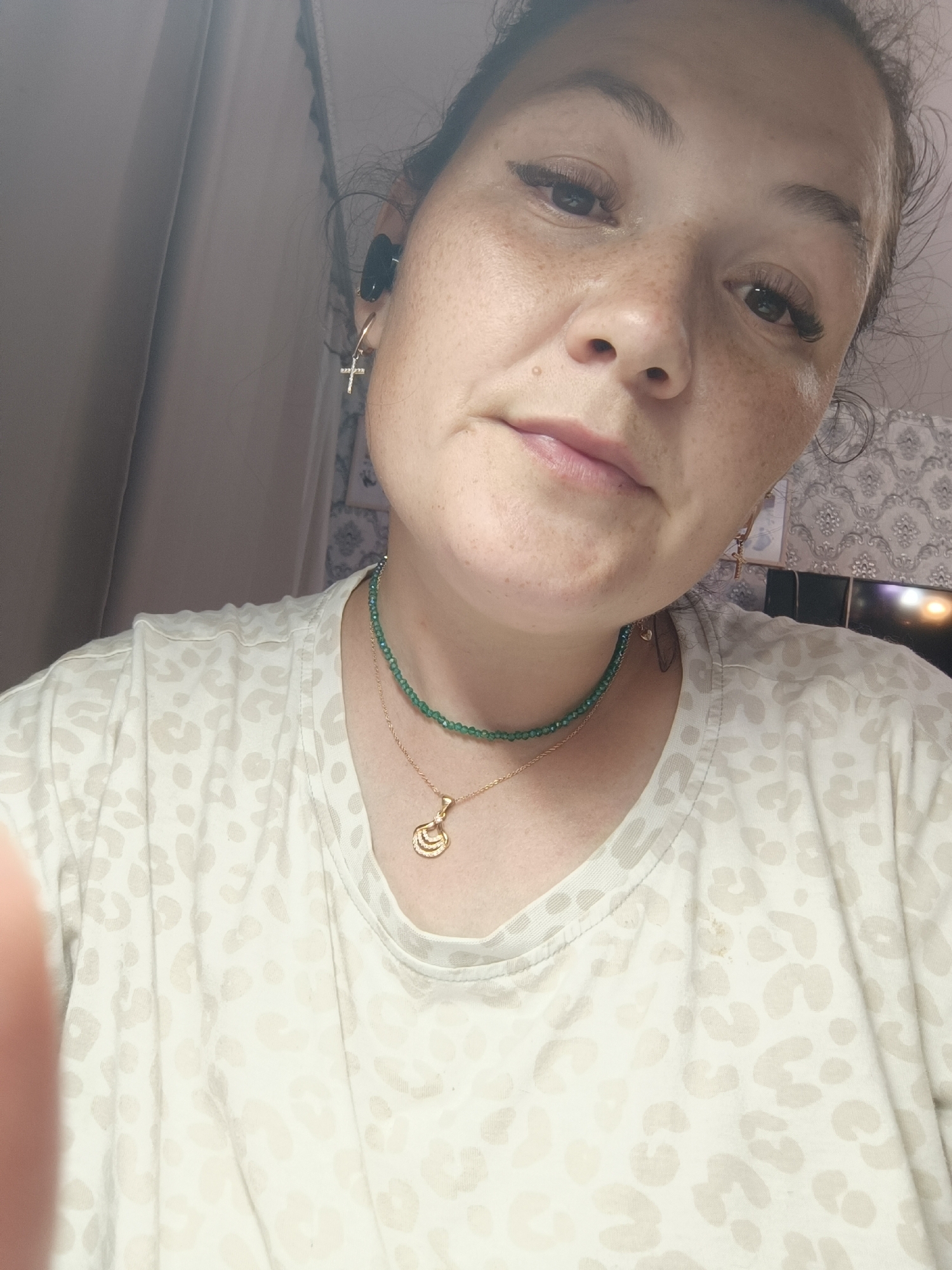 A woman is looking up at the camera. She has brown hair pulled back, freckles, and is wearing a light-colored t-shirt with a leopard print pattern. She is also wearing two necklaces: one with a green beaded string and a gold pendant, and another thinner gold chain with a pendant. She has a small cross earring in her left ear. A black earbud is visible in her right ear. The background appears to be an indoor setting with curtains and a patterned wallpaper. There is a television screen visible in the background. There is no information to confirm the location is Chaikovsky, Russia.