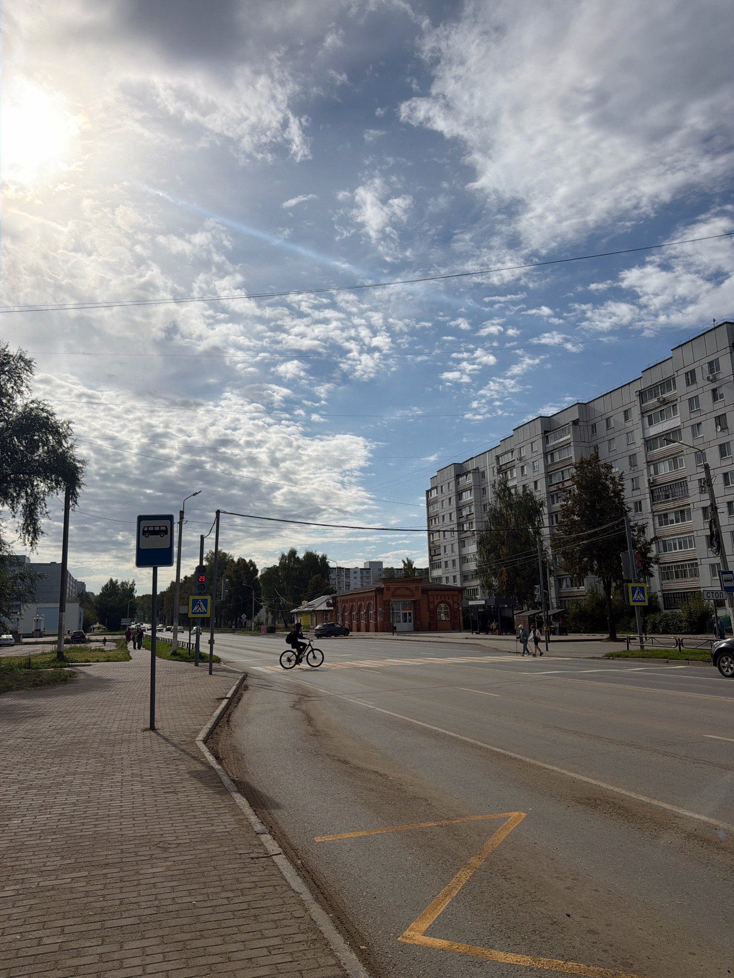 The image depicts a street scene in Chaikovsky, Russia, under a partly cloudy sky with bright sunlight illuminating the left side of the frame. A brick sidewalk runs along the left side of the road. A person is riding a bicycle across a crosswalk. Across the street, a large, multi-story apartment building dominates the background on the right. In the middle ground, a brick building with arched windows is visible, and further down the street, trees line the roadside. A bus stop sign is present on the sidewalk. A traffic light showing "23" is also visible. The road has painted lane markers and yellow markings on the asphalt. Cars are present on the street in the distance and a portion of a car is visible on the far right.