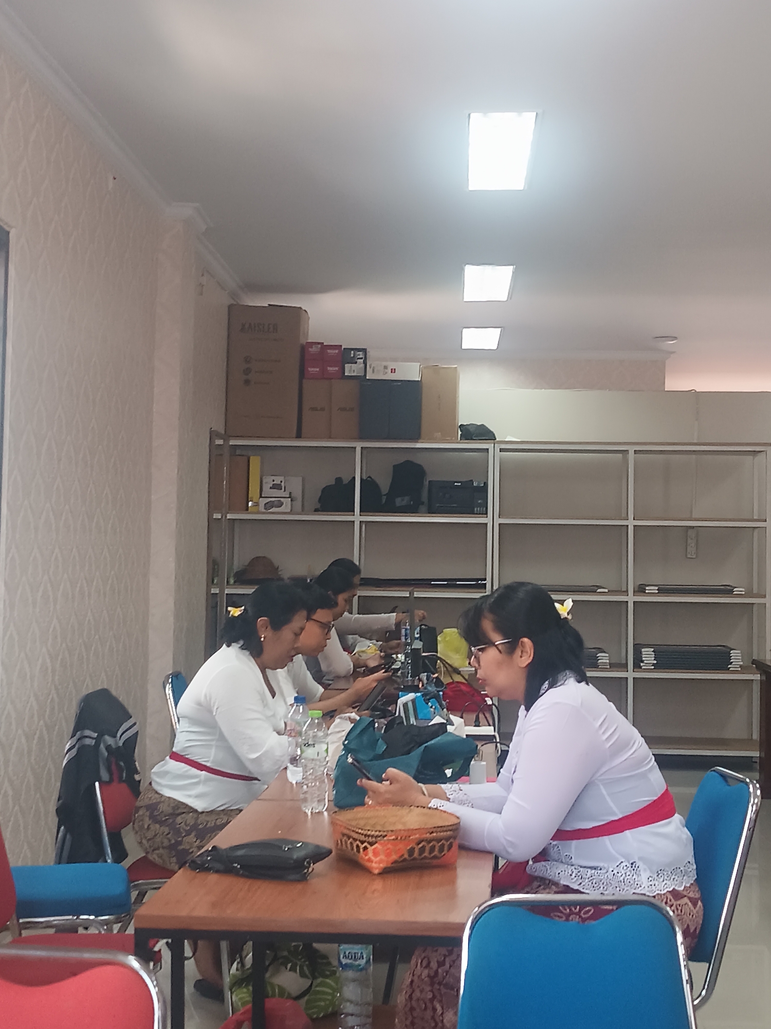 The image depicts a group of women seated at a table, engaged in what appears to be computer work. They are dressed in traditional Balinese attire, including white blouses and patterned sarongs, with red sashes tied around their waists. The environment is an indoor space with shelves filled with various items, possibly in an office or educational setting. One woman in the foreground is looking down at a laptop, while another woman next to her is also focused on a device. A third woman is partially visible in the background, seemingly engaged in a similar activity. There are bottles of water, a woven basket, and a handbag on the table. The overall scene suggests a collaborative work or learning environment within Tabanan, Indonesia, given the traditional attire.