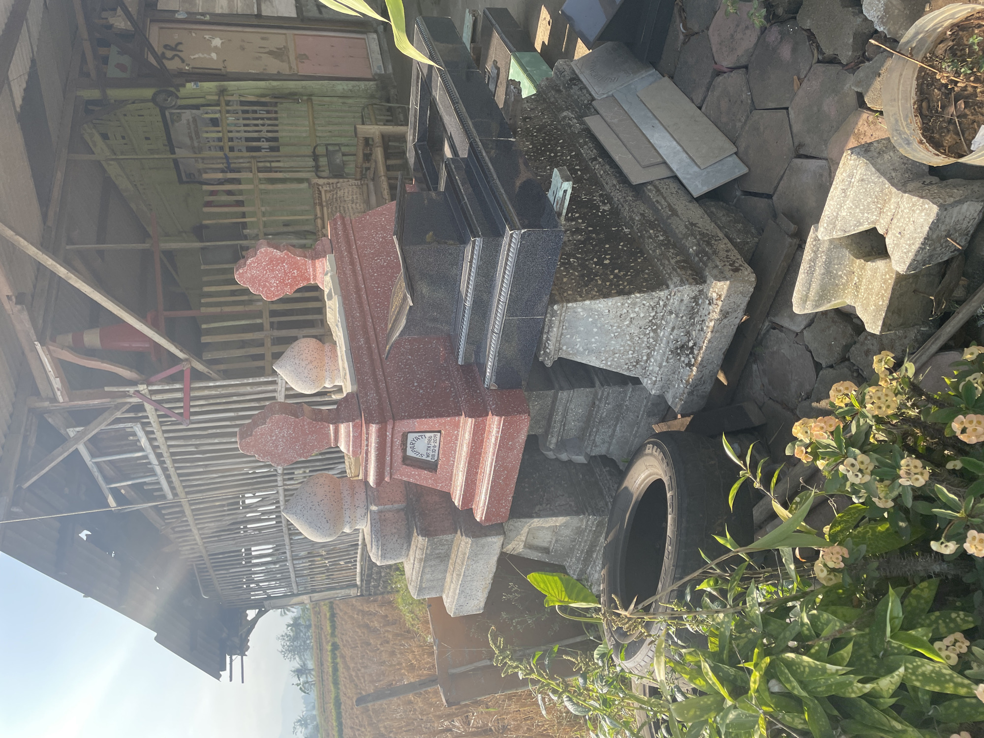 A pile of pre-made grave markers and headstones is situated in front of a rustic, open-sided bamboo structure. The grave markers are of various sizes and materials, including red and black granite. One red headstone has an inscription that reads "SUPARYAT WK TH 1996 WFB TH 1996 Dib 30-6-2014." In the foreground, there is a discarded tire, some plants with small yellow flowers, and several large, angular concrete pieces. The background shows a dry, grassy field and a distant landscape under a bright, sunny sky. The location is identified as Sucenjurutengah, Indonesia.