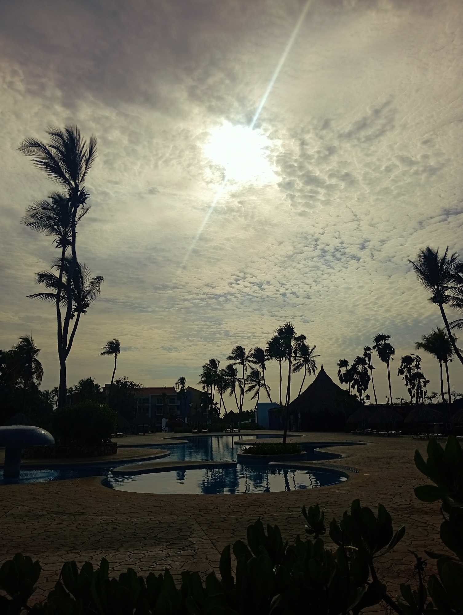 The image depicts a scene at Playa El Agua, Venezuela, showcasing a swimming pool complex with palm trees under a cloudy sky.
**Environment:** The setting appears to be a tropical resort or recreational area, given the presence of palm trees, a swimming pool, and a thatched-roof structure. The sky is filled with stratocumulus clouds, partially obscuring the sun and creating a somewhat hazy atmosphere.
**Subjects/Objects:**
* A multi-tiered swimming pool is the central element, reflecting the sky.
* Various palm trees of different heights are silhouetted against the sky, framing the scene.
* A building with a reddish roof is visible in the background, near a thatched-roof structure.
* Foliage and shrubbery are present in the foreground, adding depth.
**People:**
* There is no presence of people in the image.
**Actions/Interactions:**
* There is no presence of actions or interactions in the image.
