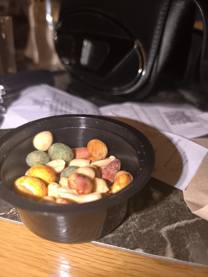 A close-up, eye-level shot prominently features a small, black plastic cup containing a variety of mixed nuts and coated snacks. The nuts display a range of colors, including natural beige peanuts and others coated in vibrant green, orange, red, and pale beige hues. The plastic cup itself is ribbed on its lower exterior and rests on a dark, patterned surface, possibly marble or stone, which in turn partially covers a lighter, wooden table surface visible in the immediate foreground.
The background is softly blurred, providing context without distracting from the main subject. Visible elements include crumpled white papers or napkins, one of which faintly shows a QR code and some unreadable text. A portion of a brown paper bag is also discernible behind the cup. To the upper right, a large, dark, out-of-focus object that appears to be an earcup of over-ear headphones or a camera lens is visible, reflecting ambient light. Another blurry, reflective object, possibly a glass, is present on the far left.
The lighting is warm and artificial, suggesting an indoor setting such as a bar, cafe, or casual dining area, likely during the evening or night. The cup casts a shadow onto the dark table surface. There are no people visible, implying the snacks are prepared for consumption. While the location context is Palaio Faliro, Greece, there are no specific visual cues within the image to confirm this.