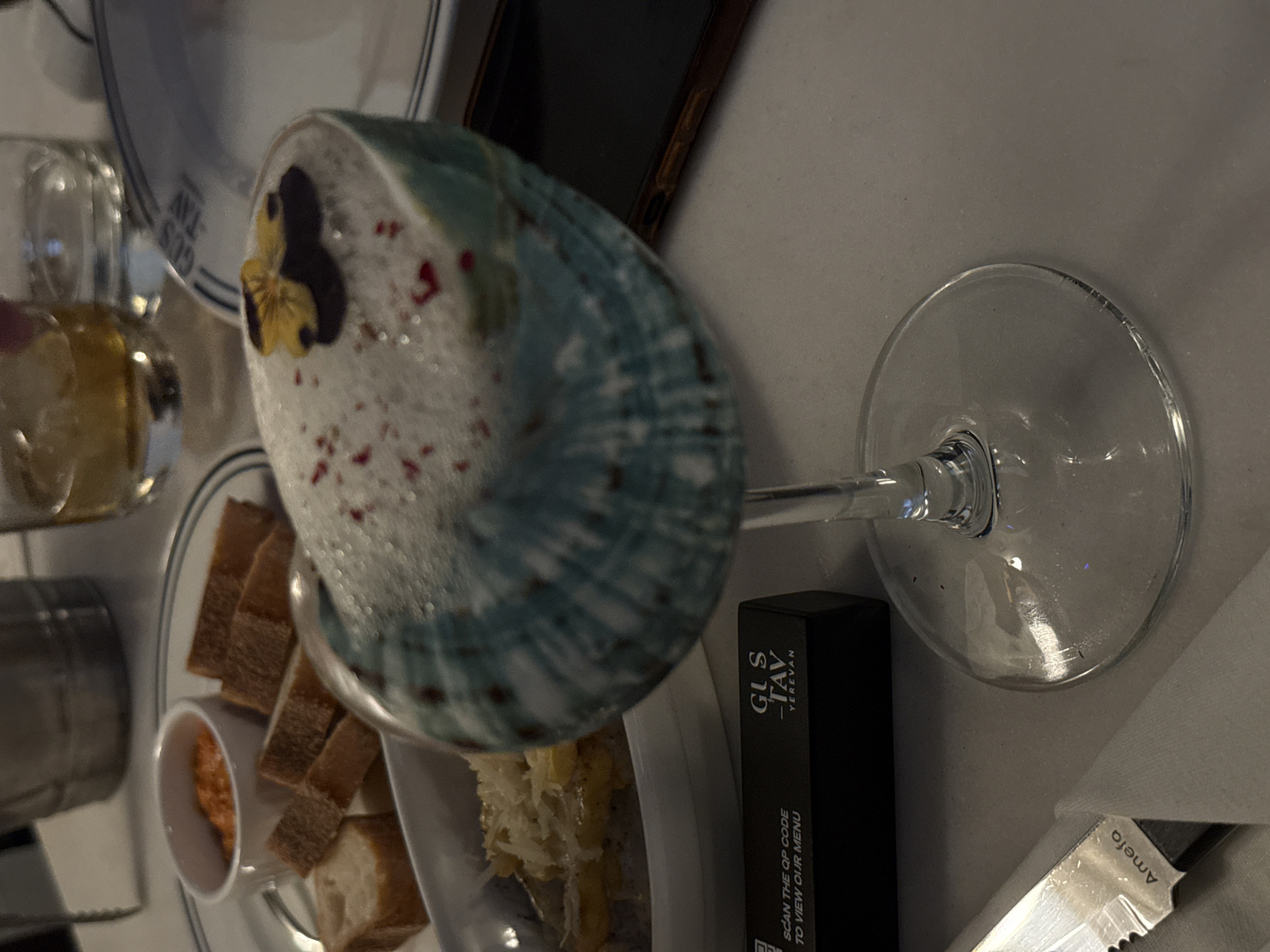 The image shows a close-up of a table setting, likely in a restaurant in Yerevan, Armenia. The main subject is a dessert served in an iridescent, seashell-like dish, topped with a pansy flower and red sprinkles. Beside the dessert, there are pieces of bread and a small ramekin of what appears to be a sauce or spread. A wine glass with its base to the right of the image is also visible, reflecting light. A black menu holder with the text "GUS - TAV YEREVAN" and "SCAN THE QR CODE TO VIEW OUR MENU" is positioned near the wine glass. A knife with "Amefa" written on it is also present on the table. The setting appears to be indoors, and the lighting suggests it might be evening. The overall impression is of a fine dining experience.