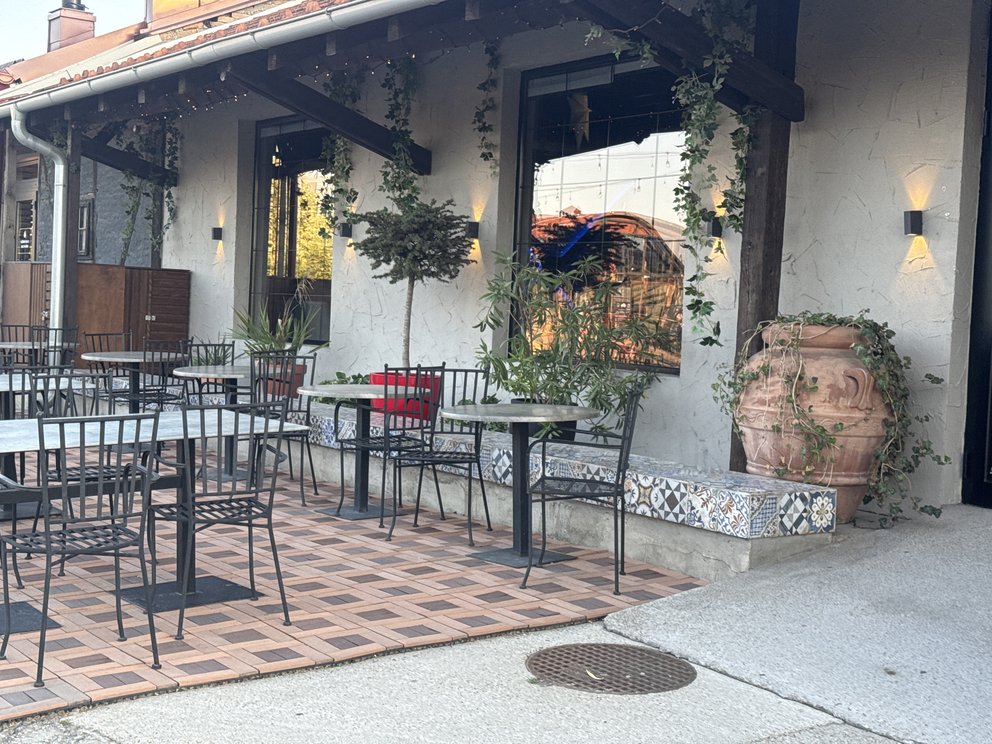 This image depicts the outdoor seating area of a restaurant or cafe, located in Belgrade, Serbia. The scene is characterized by an arrangement of metal tables and chairs on a patterned tiled floor. In the background, the building features large windows reflecting an outdoor scene, possibly a street or another building with colorful lights. A large terracotta pot with trailing ivy and a decorative tiled bench are also prominent features of the outdoor decor. The overall ambiance suggests a pleasant, possibly evening, setting for dining or socializing.