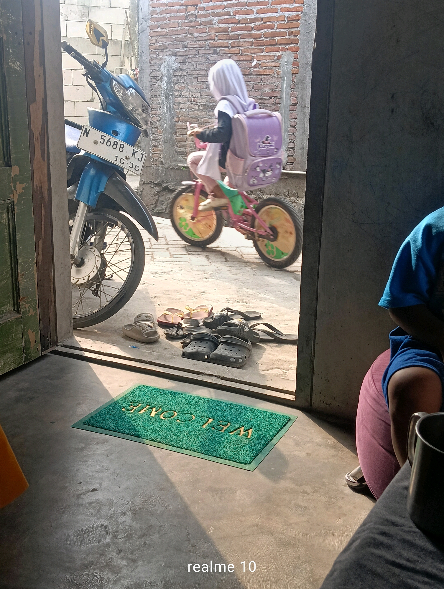 In Batu, Indonesia, an outdoor scene captured on a bright, sunny day features a young child, seen from behind and the side, wearing a white hijab and a pink dress, riding a bicycle. The child carries a light purple backpack with a duck design. To the left, a blue motorcycle with a license plate reading "N 5688 KJ 10.JG" is parked. The ground is concrete, with several pairs of sandals and flip-flops scattered near the motorcycle. A green welcome mat with yellow lettering is placed on the floor in the foreground, partially illuminated by sunlight. To the right, a person wearing a blue shirt and pink pants is partially visible, seated on a red cushion. The background consists of brick walls.