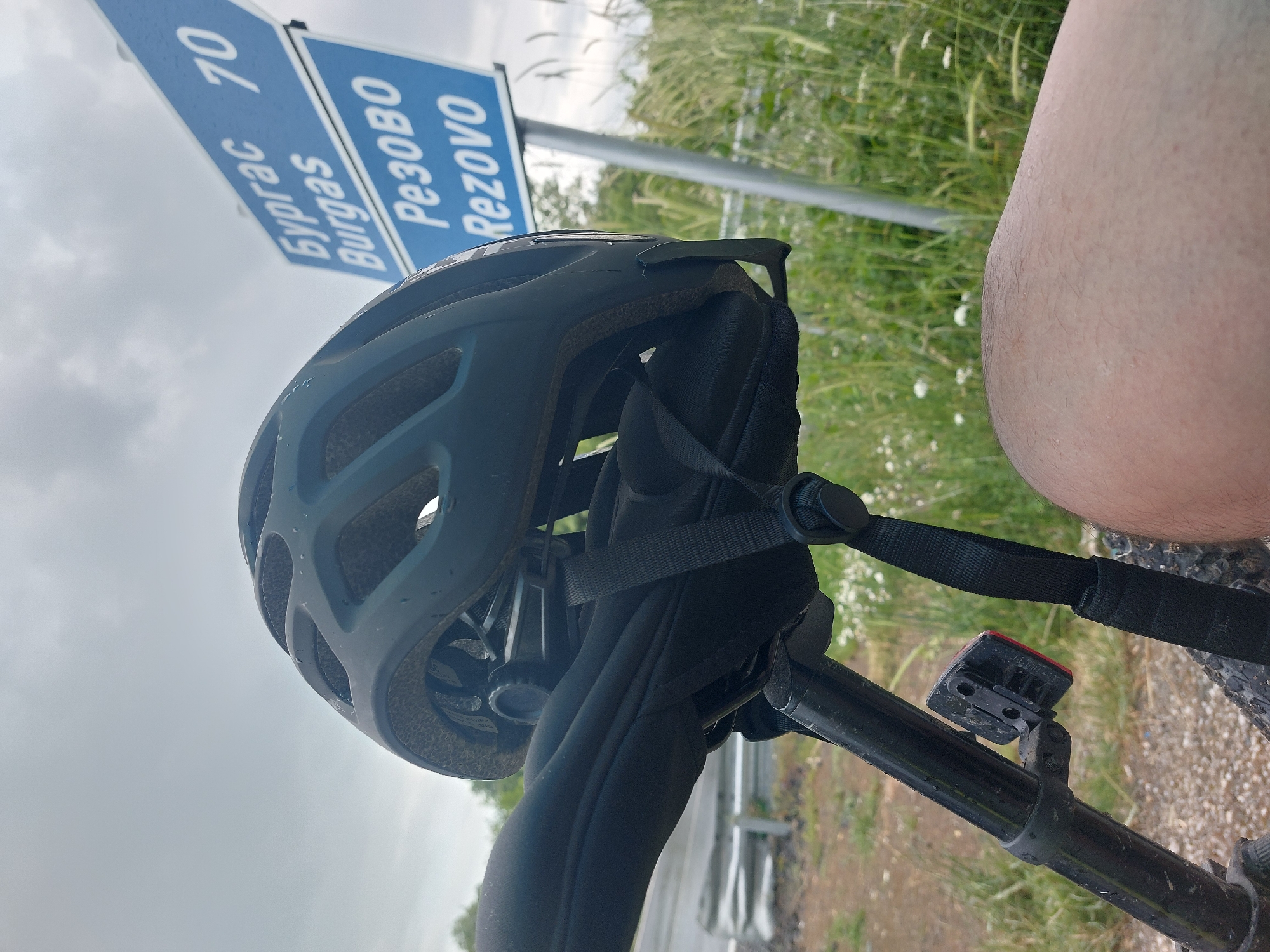 A bicycle helmet rests on the bicycle handlebar, a road sign indicating "Burgas 70, Rezovo" visible in the background. The location is likely near the city of Tsarevo, Bulgaria, as Rezovo is a village in the Tsarevo Municipality. A leg is partially visible, and a small red light is mounted on the handlebar. The weather appears overcast.