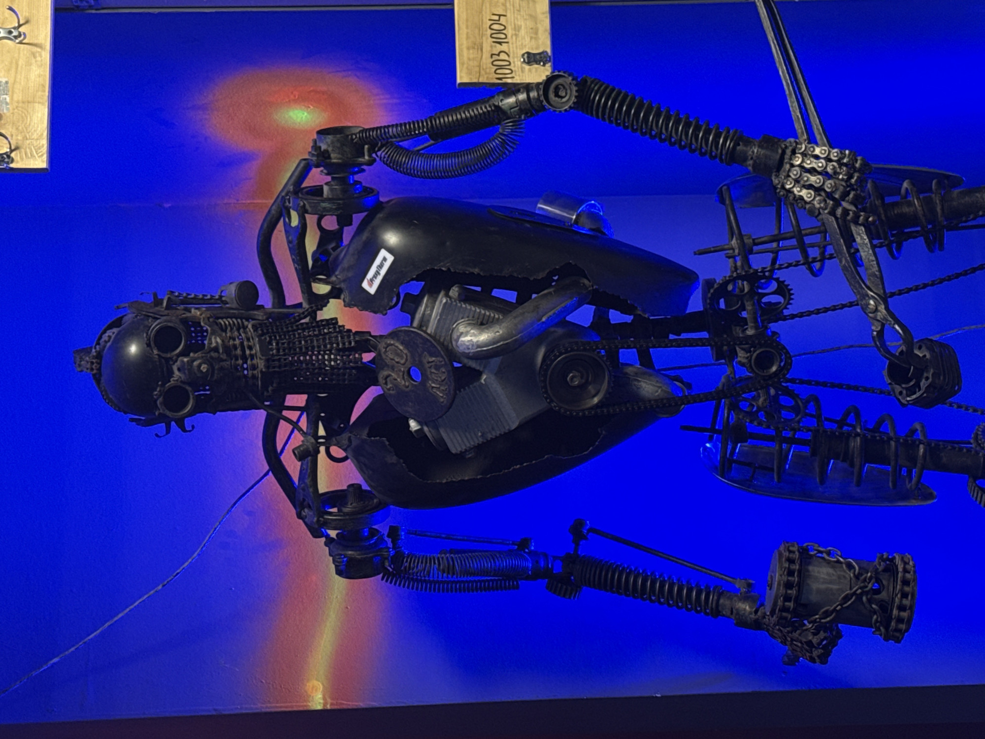 The image shows a full-body sculpture of a robot made from recycled metal parts, displayed against a vibrant blue background with red and yellow light projections. The robot has a metallic skull-like head with goggles, a segmented chest cavity revealing inner mechanisms, and articulated arms and legs. The sculpture appears to be an exhibit, possibly in a museum or gallery. There is a wooden sign with the numbers "1003 1004" to the right of the robot. The robot's materials suggest it's a piece of industrial art. No people are visible in the image. The background lighting creates a dramatic effect, highlighting the metallic textures of the robot.